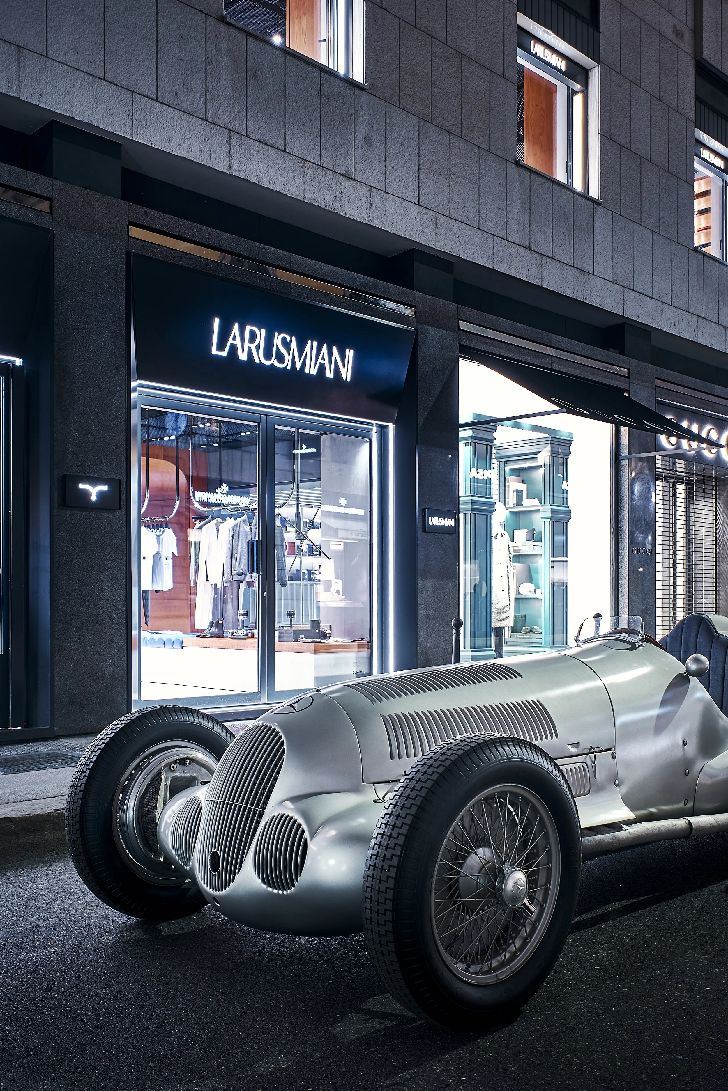 A vintage silver race car parked outside a luxury clothing store named Larusmiani, at night.