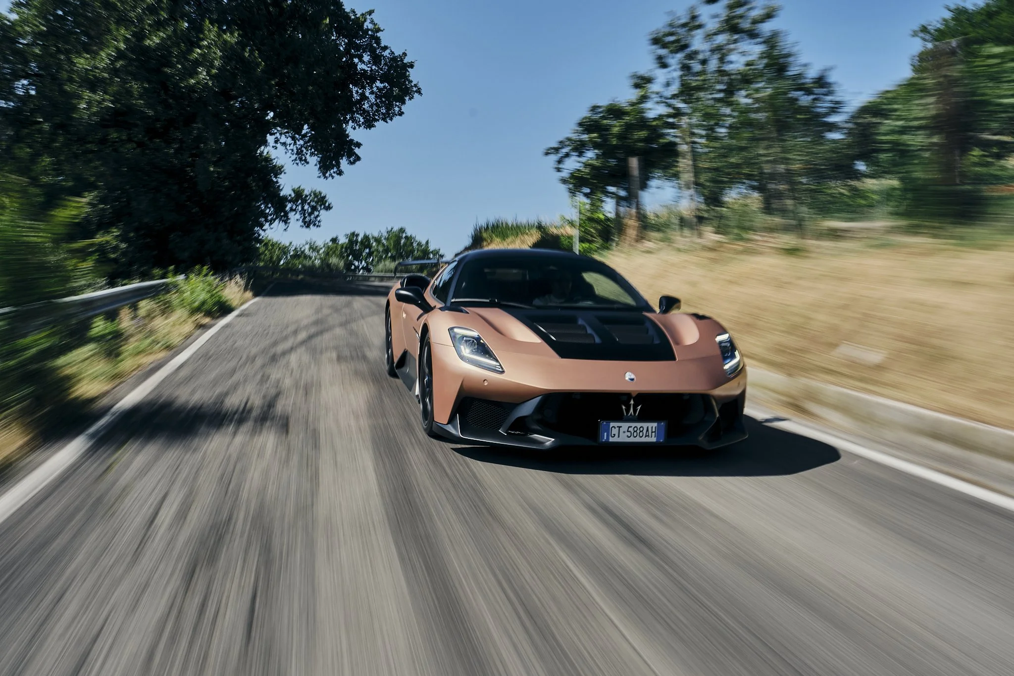 A tan and black Maserati sports car driving on a curvy mountain road with trees on either side.