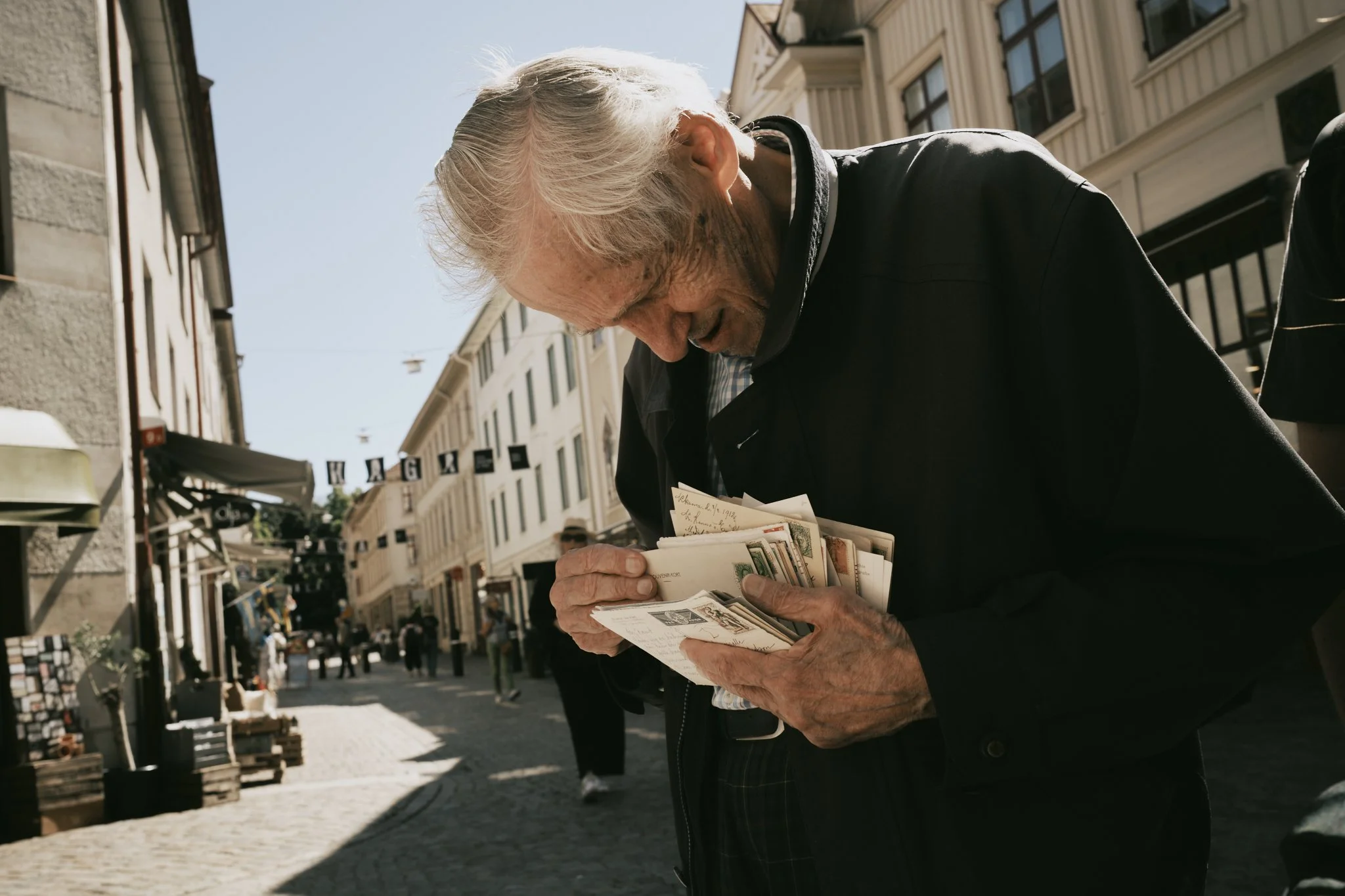 An elderly man with white hair and a black jacket looking at postcards or photographs on a street in a city, with buildings and pedestrians in the background.