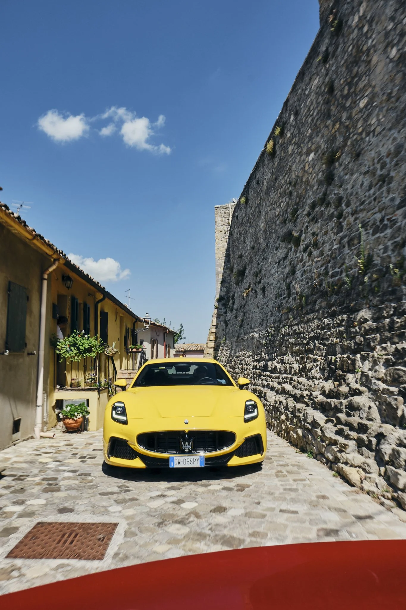 A yellow Maserati car parked on a cobblestone street beside a stone wall in a quaint town with colorful houses and potted plants under a blue sky.