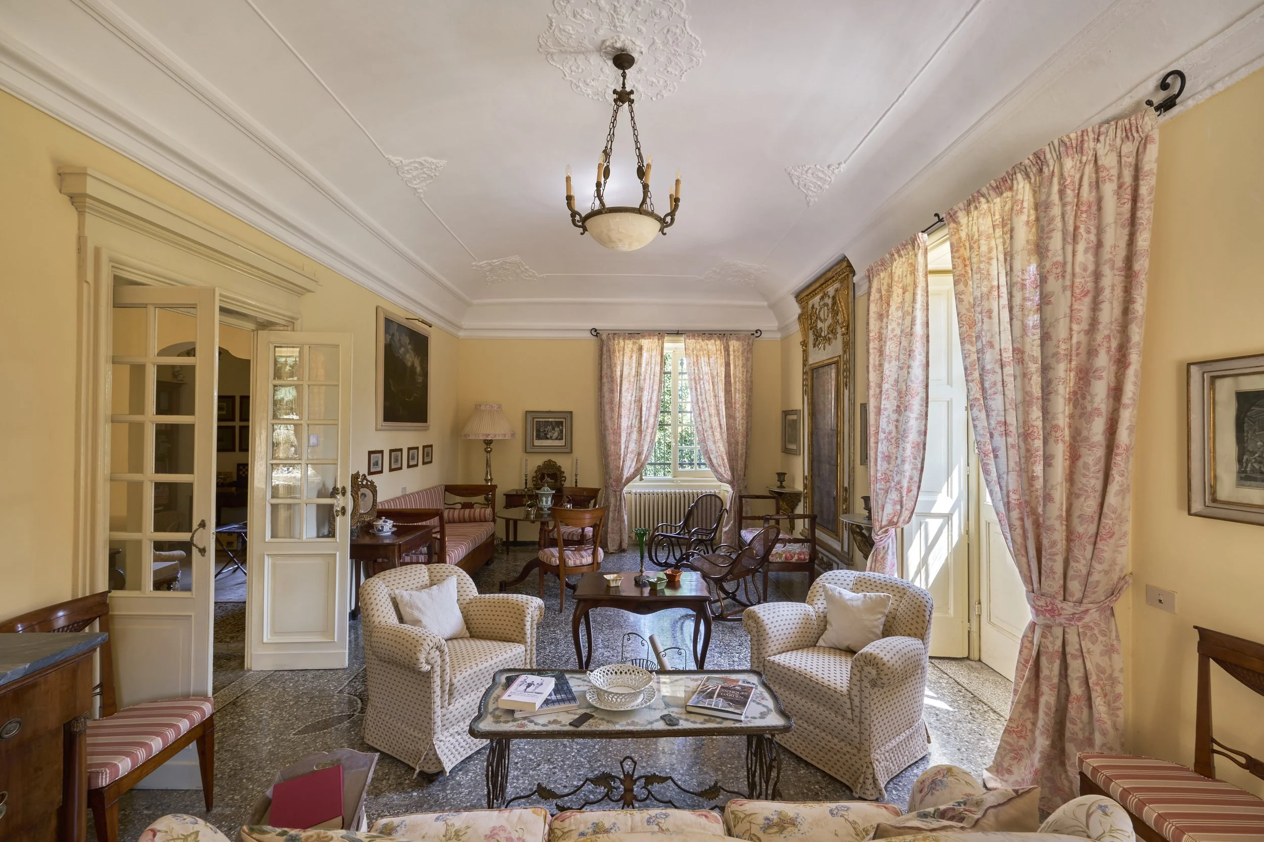 Interior of a vintage living room with yellow walls, ornate ceiling molding, pink curtains, antique furniture, and a chandelier.