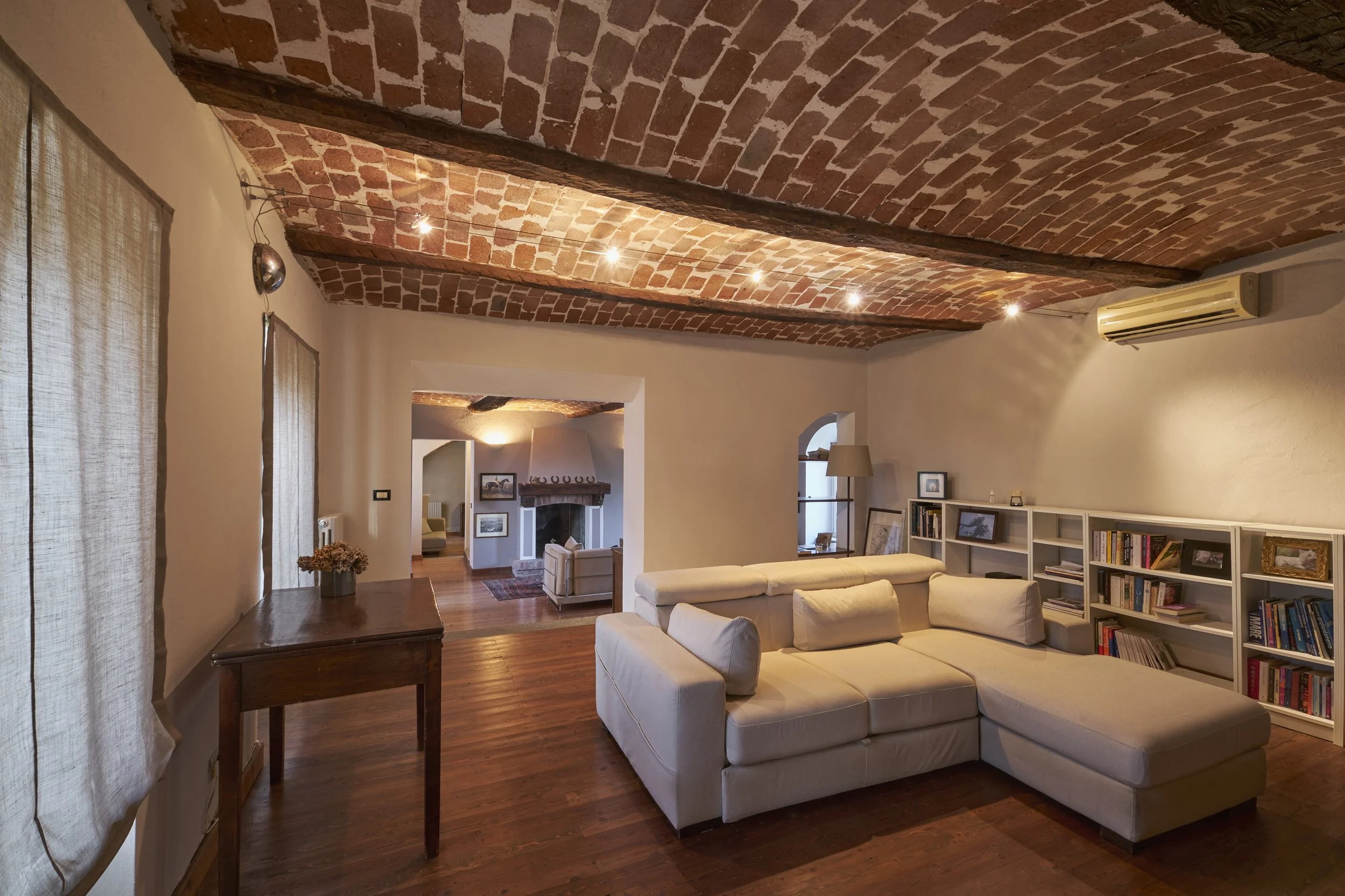 Living room with brick vaulted ceiling, white sectional sofa, wooden floors, bookshelf, and a fireplace in the background.