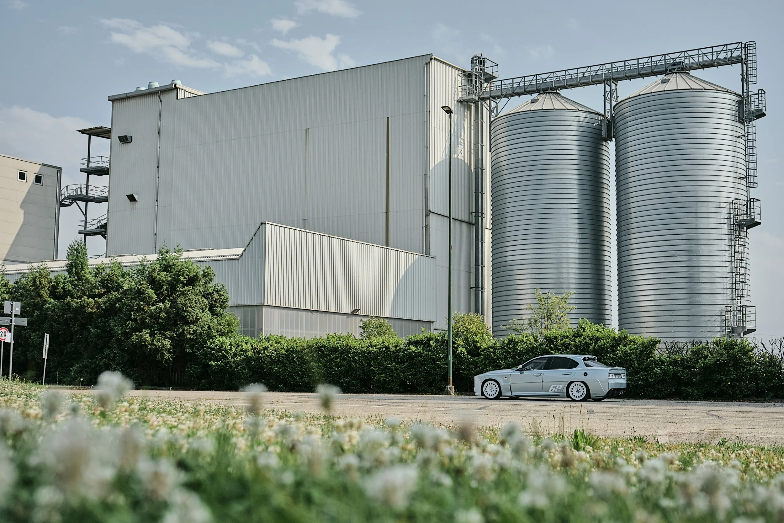 Industrial building with two large silos, parked silver sports car in front, greenery and flowers in the foreground, blue sky with some clouds.