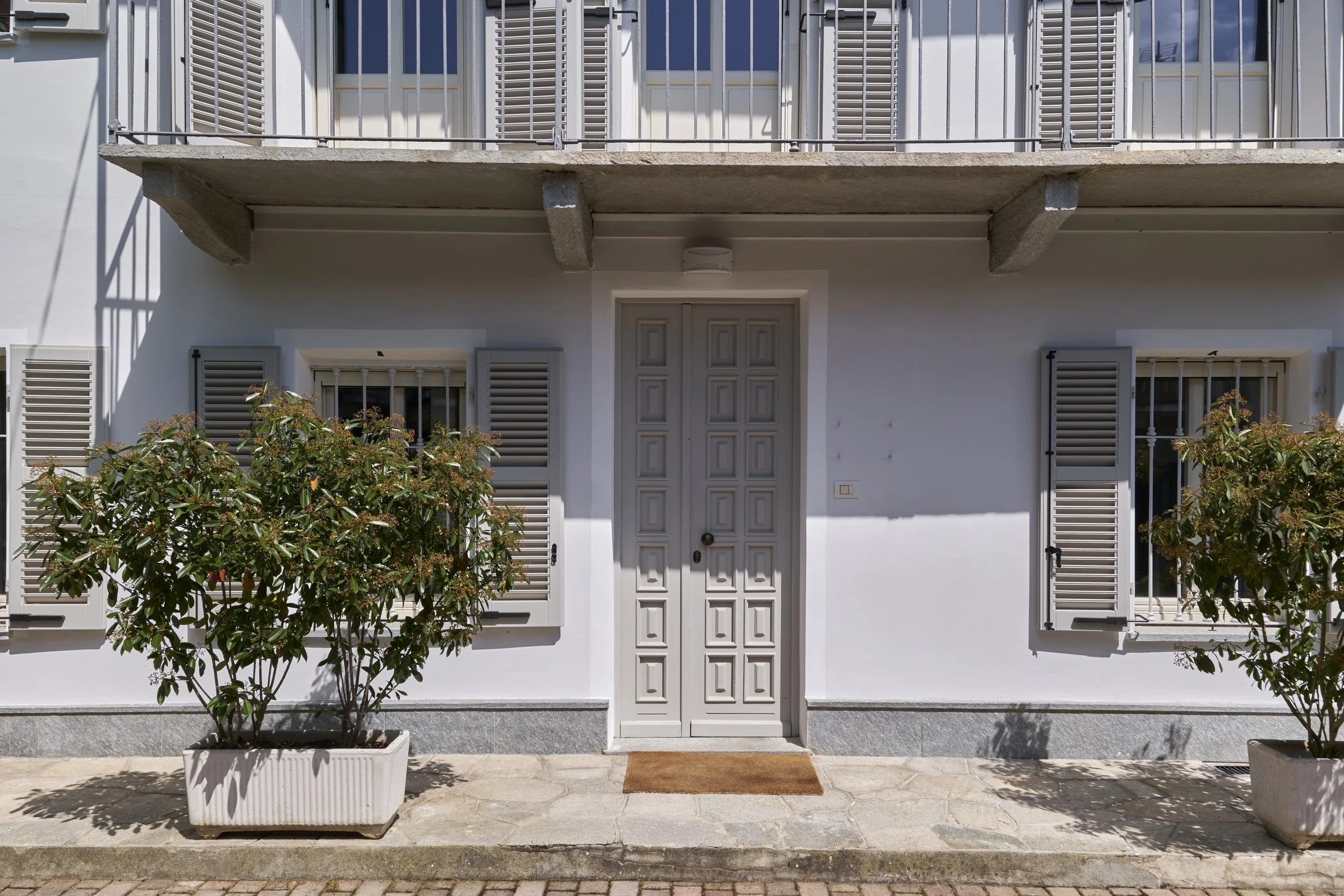 White building entrance with a double door, two windows with shutters, potted plants, and a small rectangular doormat