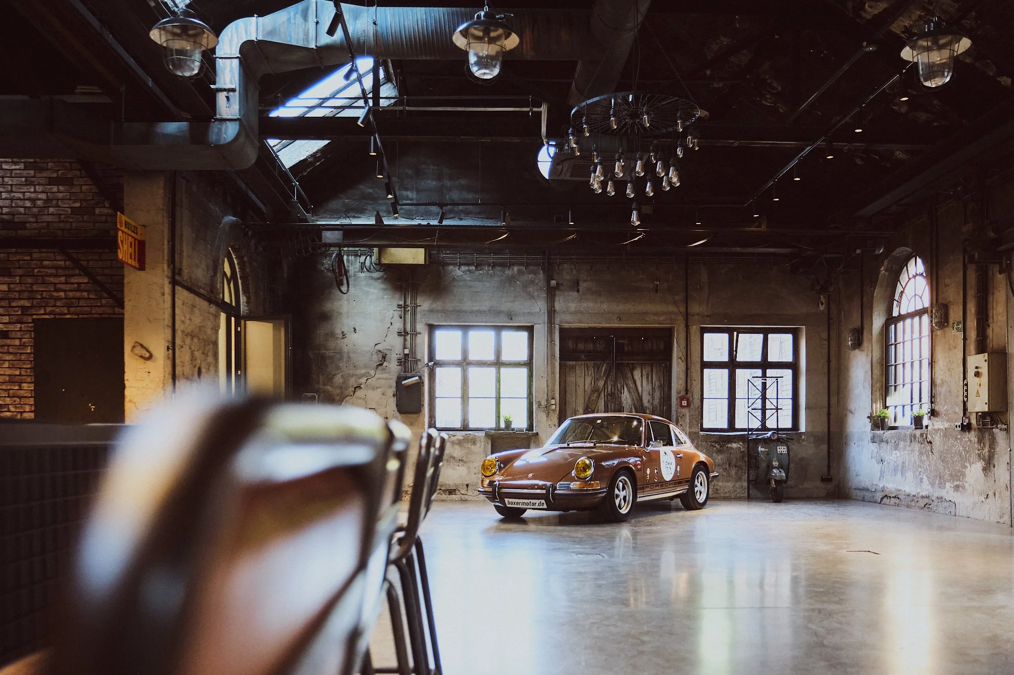 A vintage red Porsche car parked indoors in an industrial-style space with large windows, exposed brick walls, and a chandelier hanging from the ceiling.