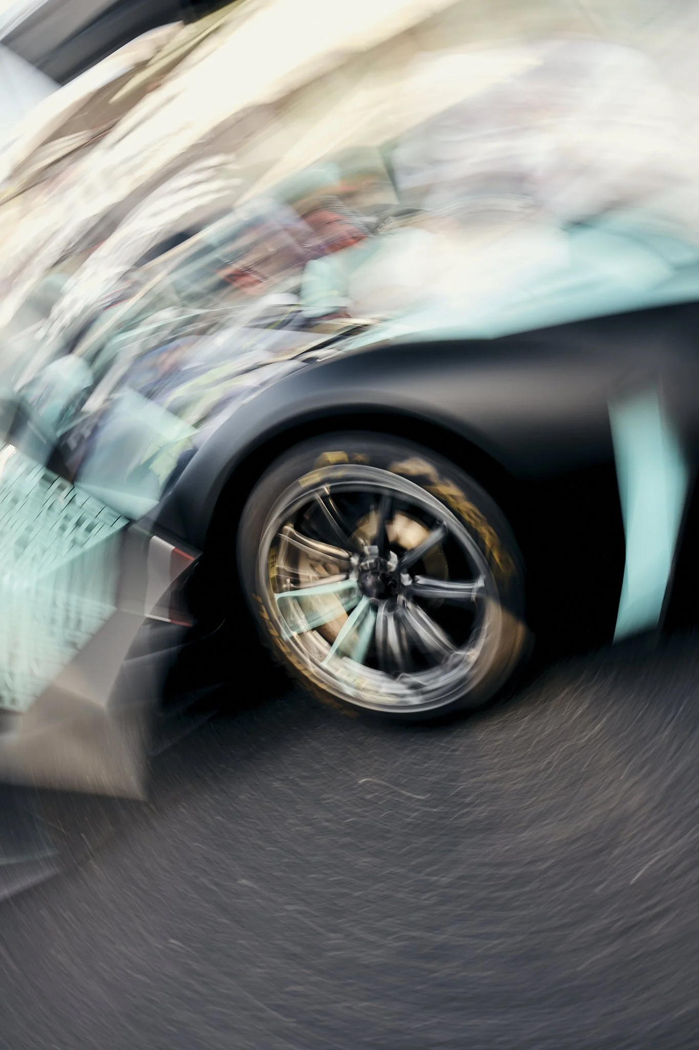Blurred photo of a sports car with a black and turquoise color scheme, showing the front wheel and part of the car's body.
