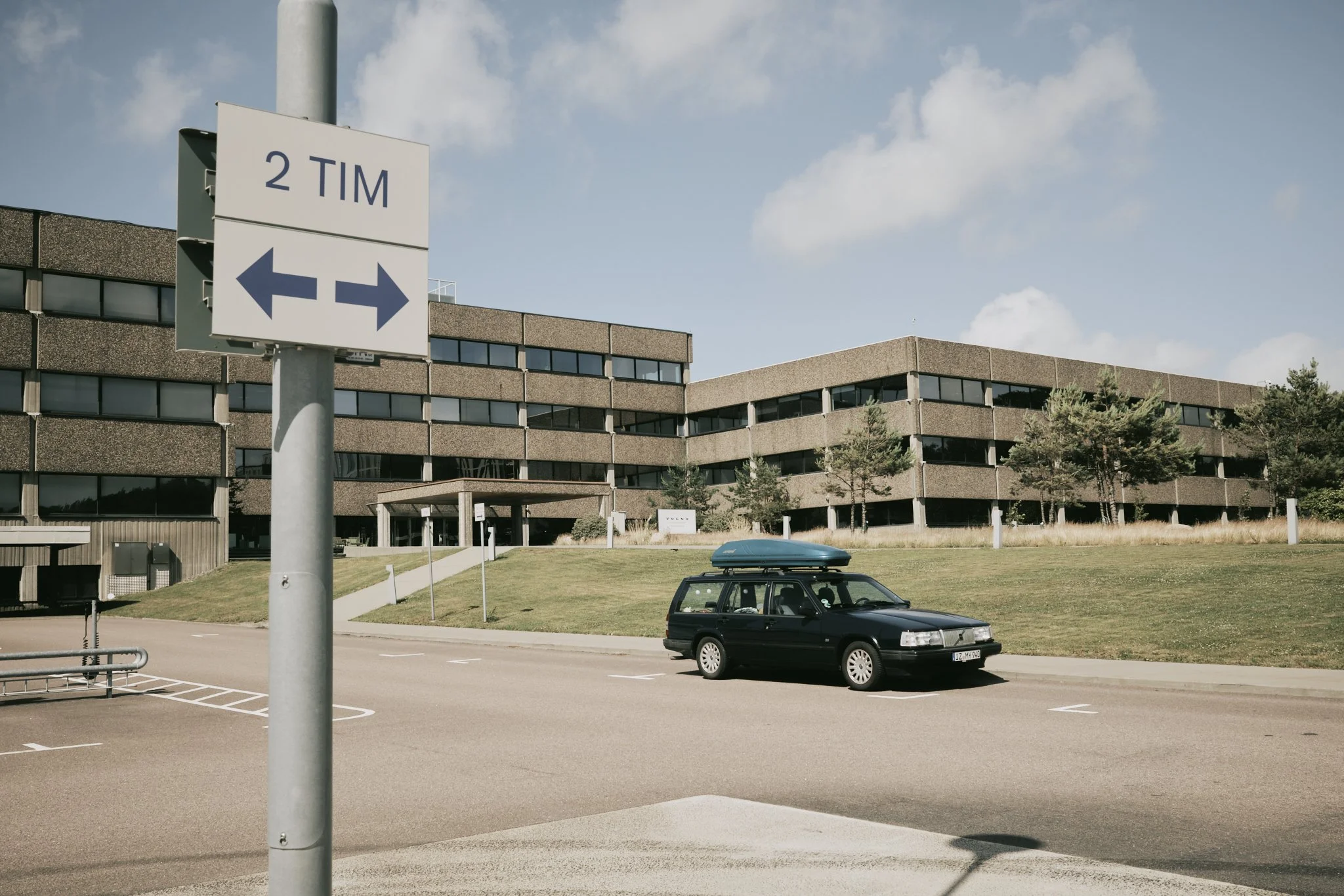 A black station wagon with a blue cargo box on top parked on an empty street in front of a modern office building, with a sign showing directions for tram line 2.