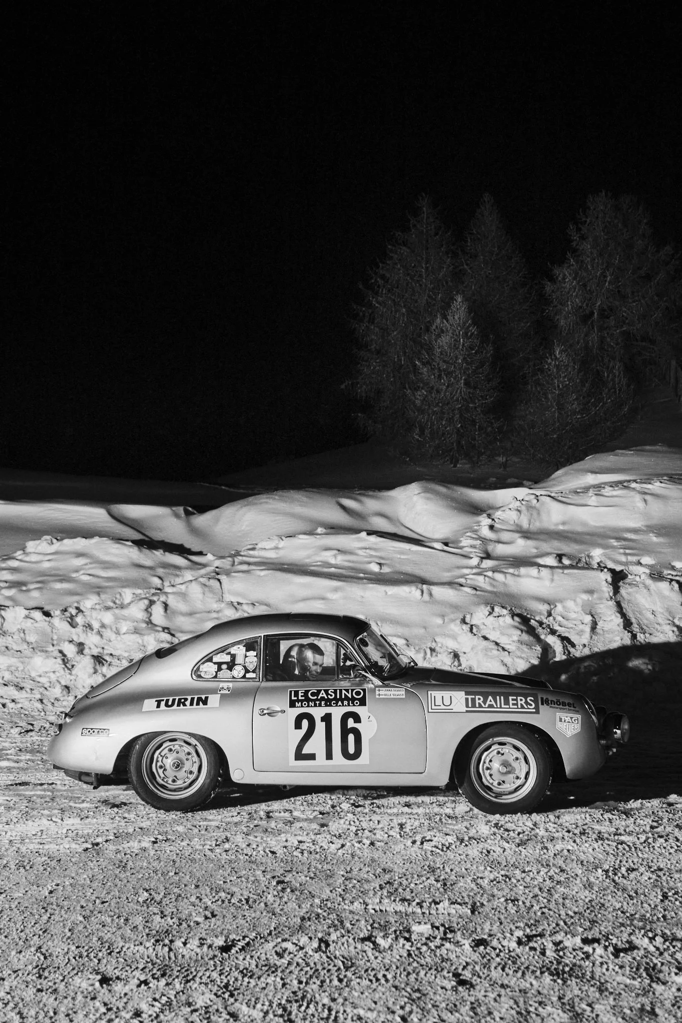 A vintage race car with the number 216 parked on a snowy road at night, with trees and snow-covered ground visible in the background.