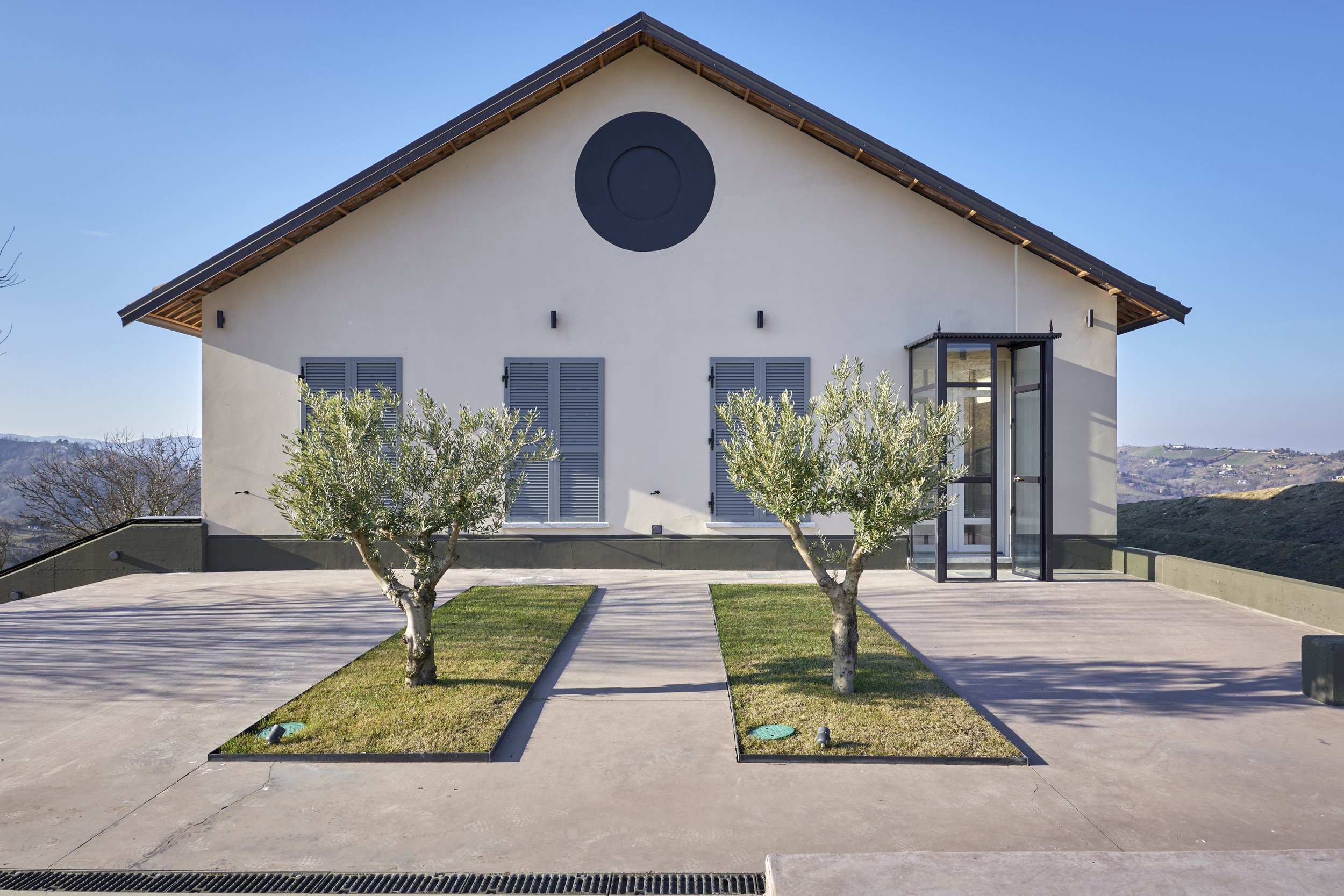 A modern white house with a sloped roof, blue window shutters, and a glass enclosed porch, surrounded by a concrete driveway and two small trees in grass patches.