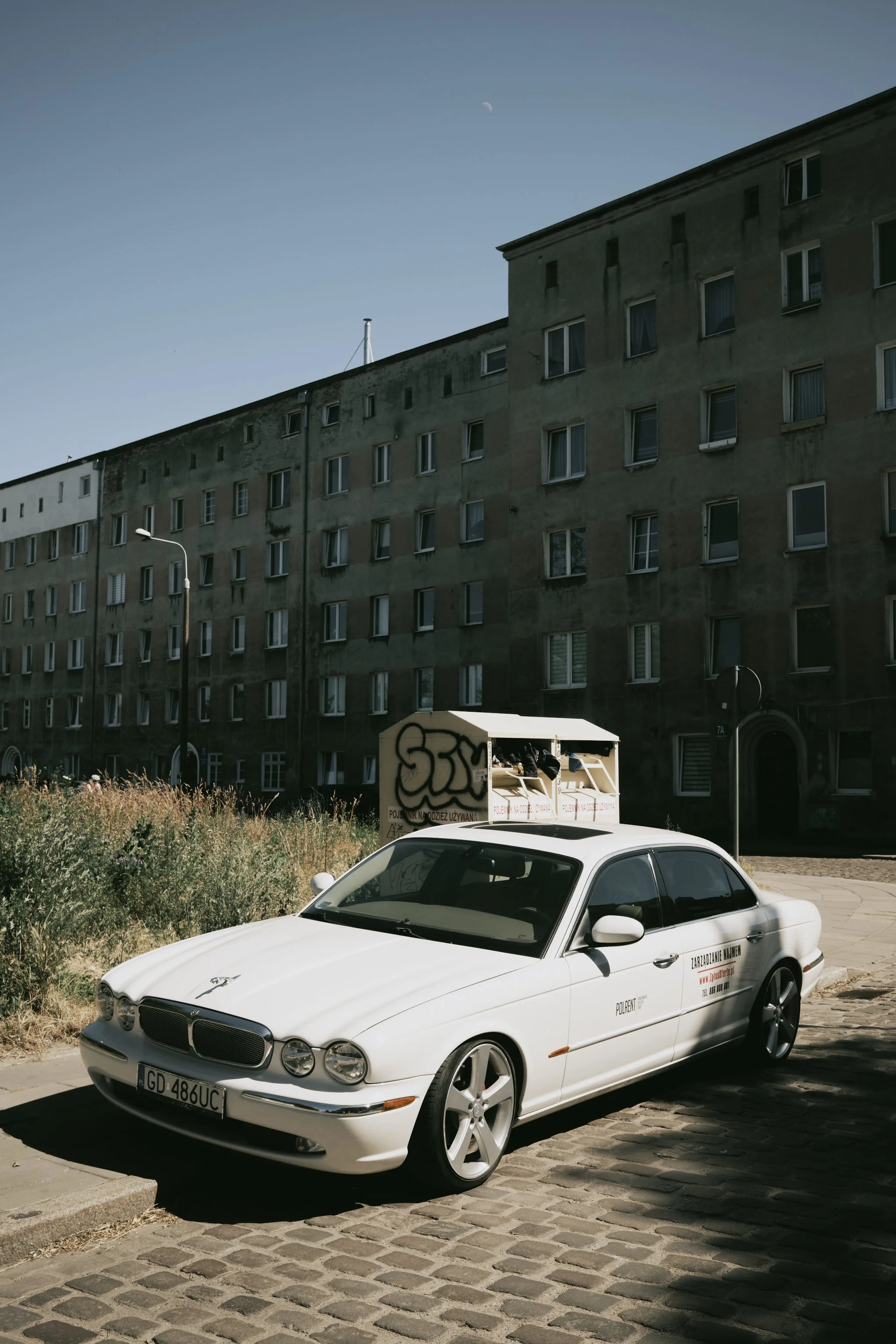 A white luxury sedan parked on a cobblestone street with an urban building in the background and graffiti on a nearby utility box.