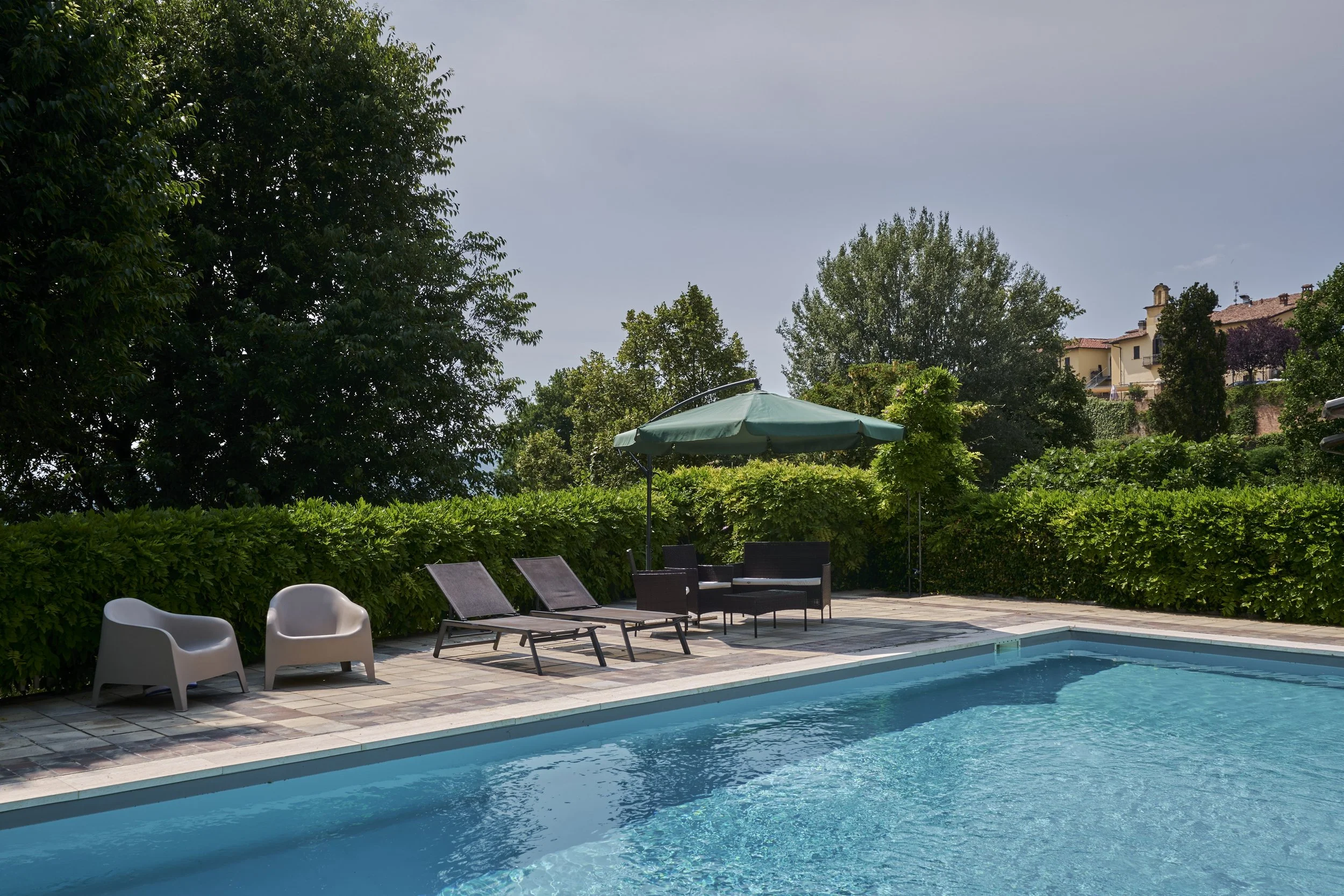 A backyard swimming pool with lounge chairs, a green umbrella, and surrounding greenery in a sunny setting.
