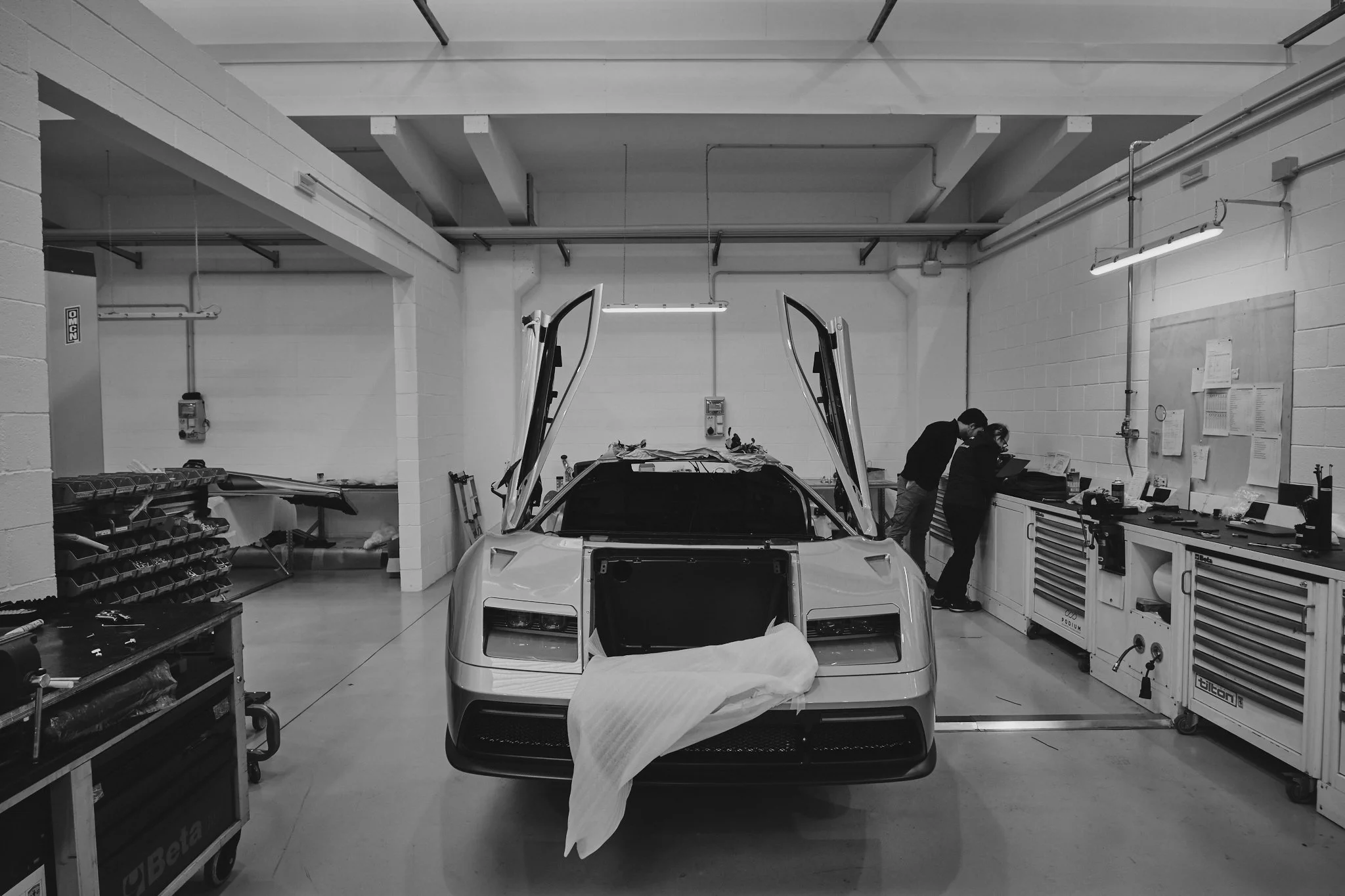 A sports car with butterfly doors open in a workshop, with two people inspecting or working on it.