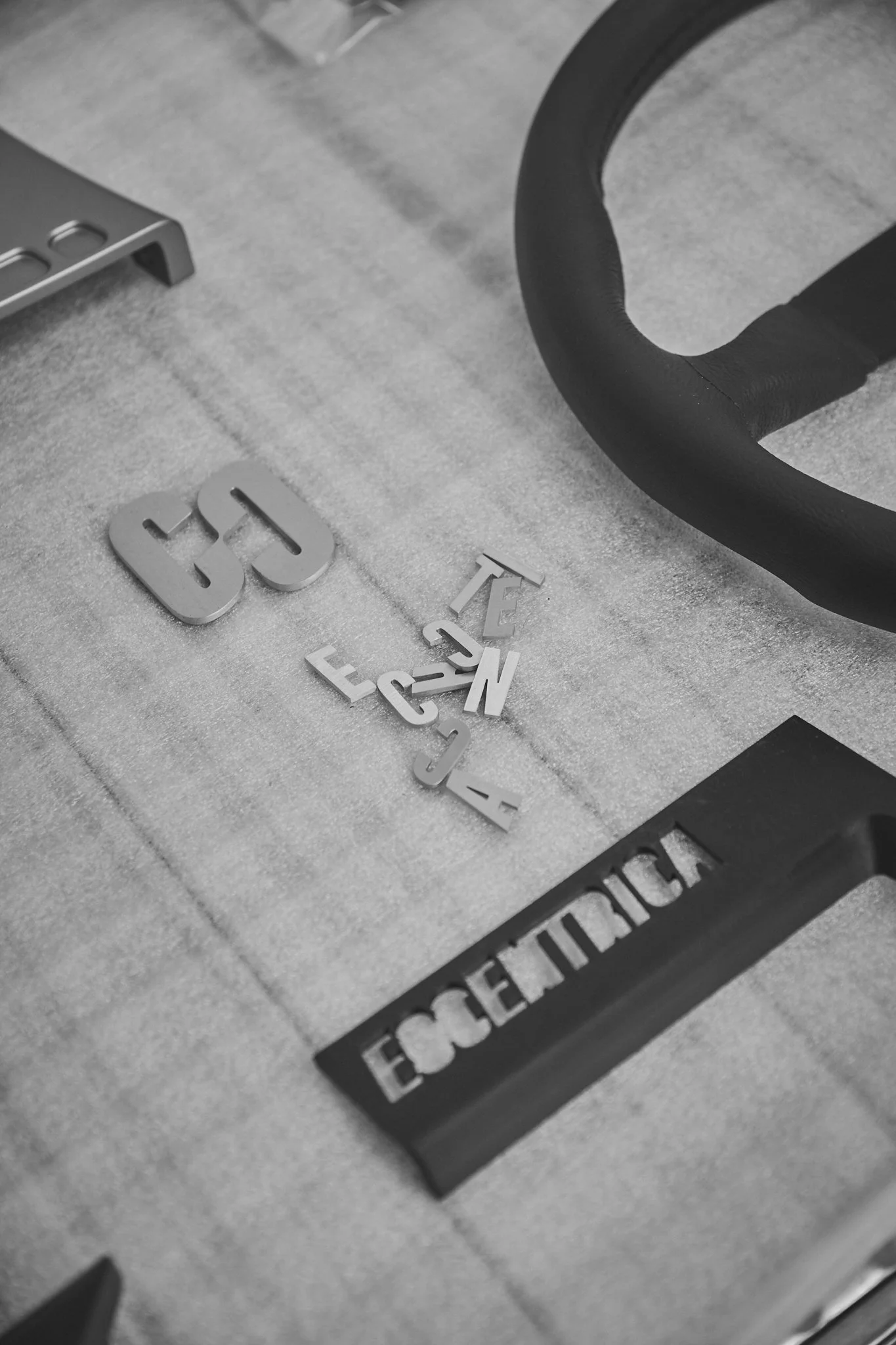 Black and white photo of scattered letter and number pieces on a surface, with a partially visible weightlifting bar and a grip strap labeled 'ENCEDRICO'.