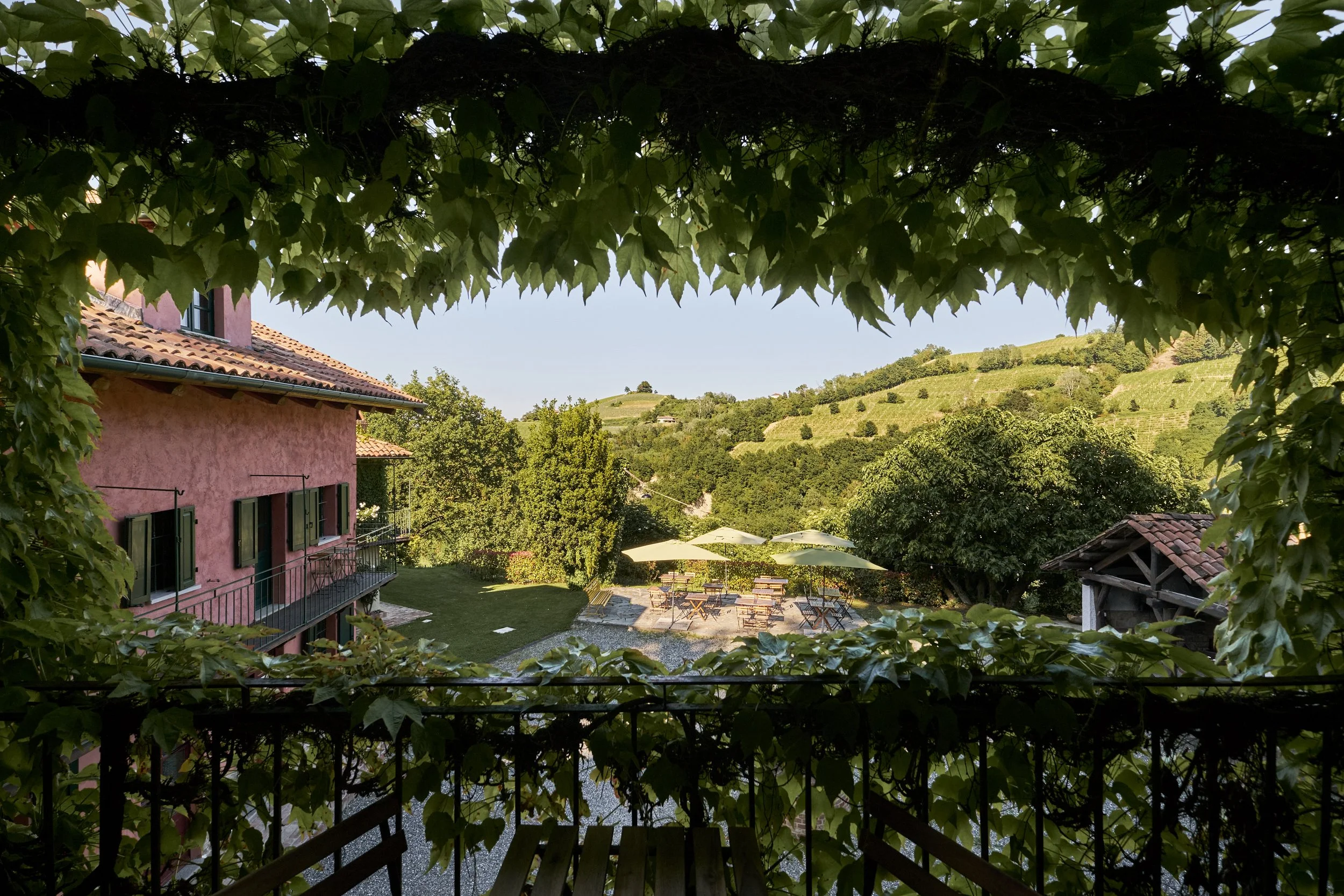 A view of a pink house with green shutters, a balcony, and a garden with outdoor seating, umbrellas, and lush green trees and rolling hills in the background, seen through a leafy frame.