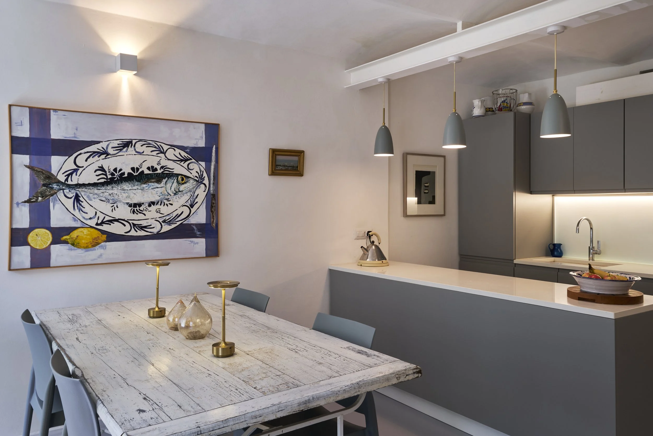 Modern kitchen and dining area with artwork depicting a fish on the wall, white table with three candlesticks and glass vases, gray cabinets, pendant lights, and a bowl of fruit on the counter.