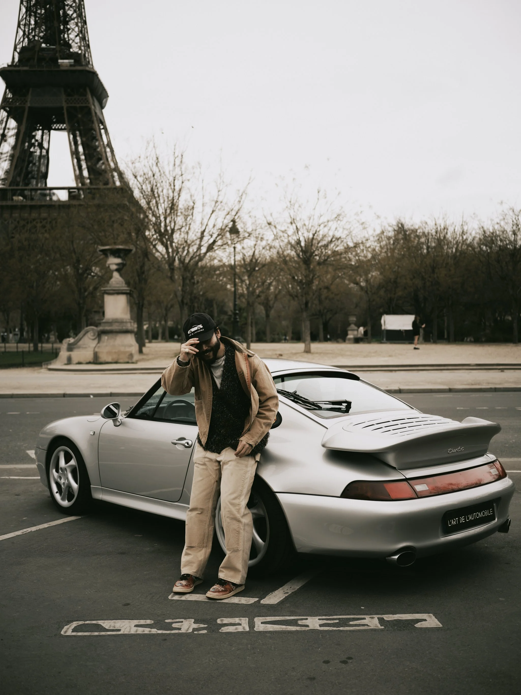 A man with long hair and sunglasses leaning against a silver Porsche sports car parked on the street, with the Eiffel Tower visible in the background.