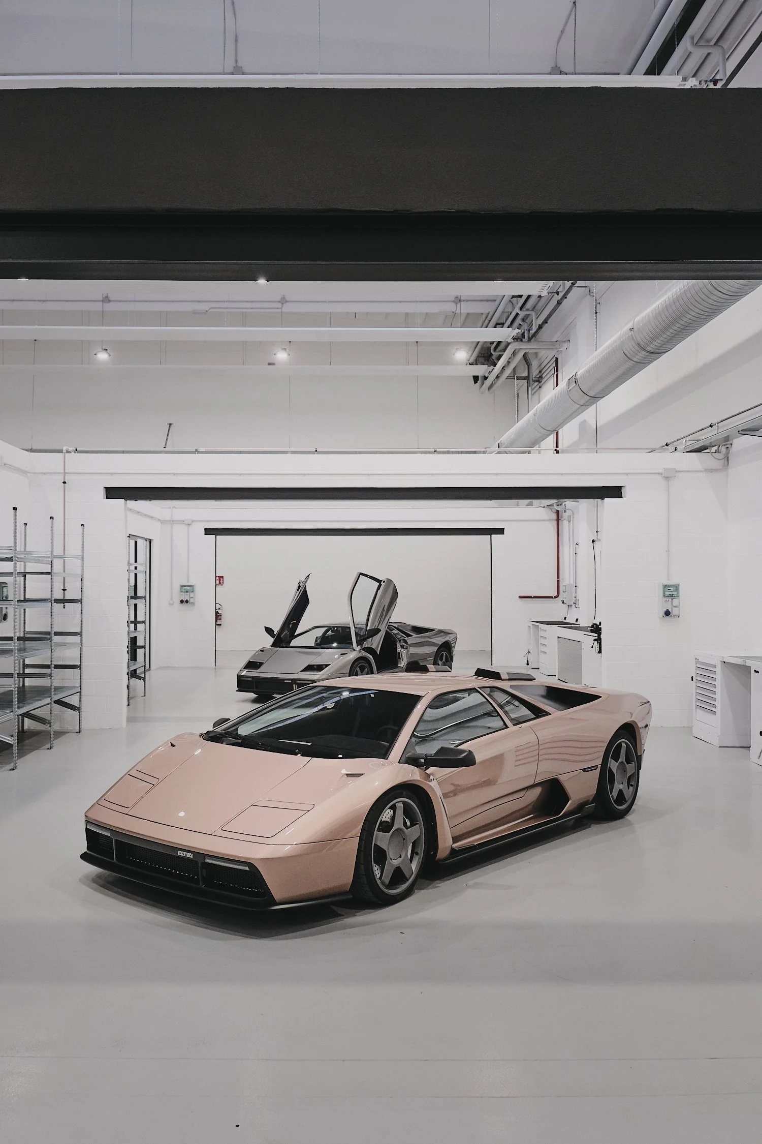 Two luxury sports cars in a clean, white garage with overhead lighting. The car in the foreground is a low, pinkish-beige supercar, and the car in the background is a gray sports car with its doors open.