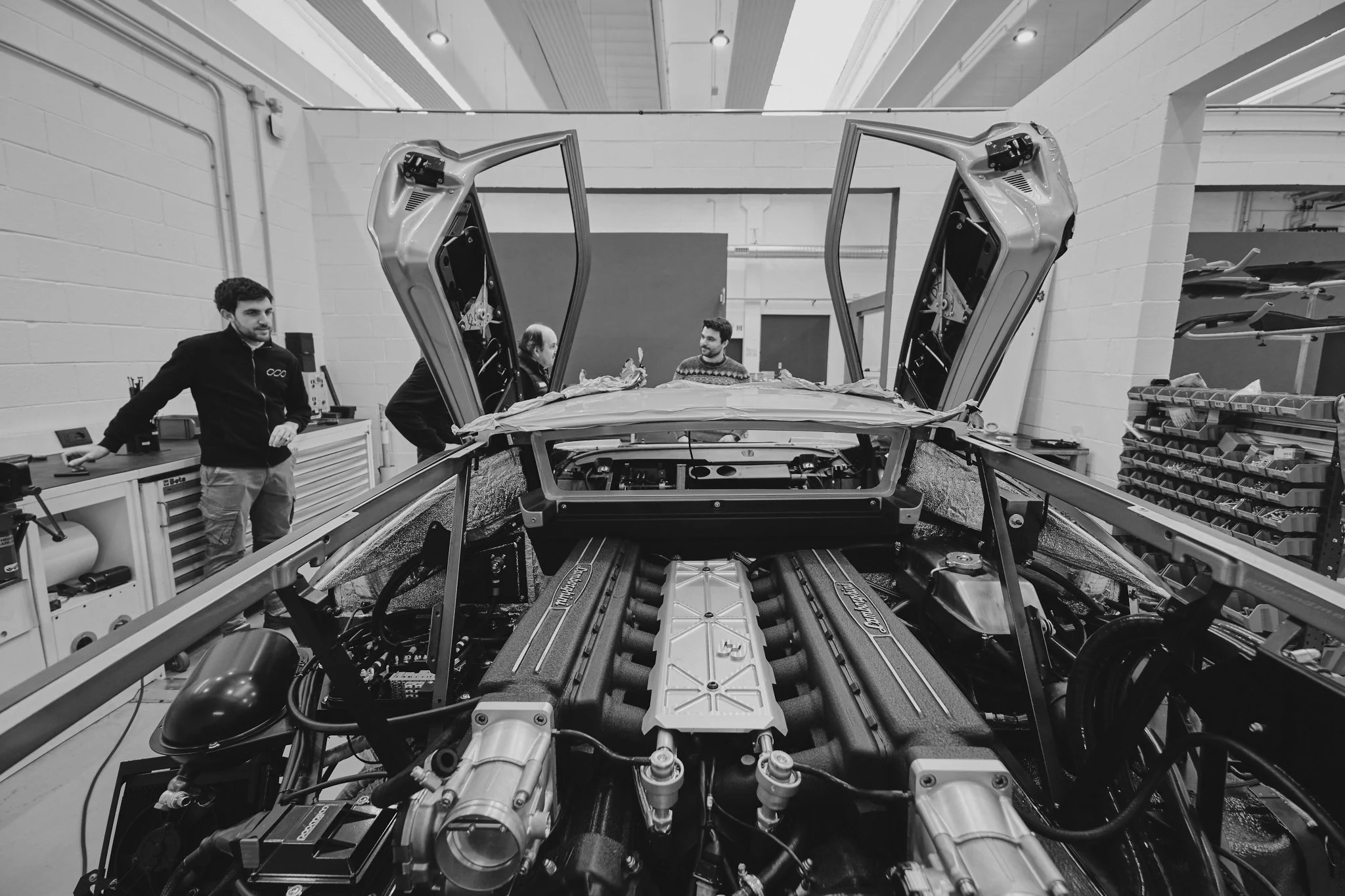 Black and white photo of a high-performance car being built or repaired in a workshop, with three men inspecting the car from behind and beside it. The car's doors are open vertically, revealing the interior framework.