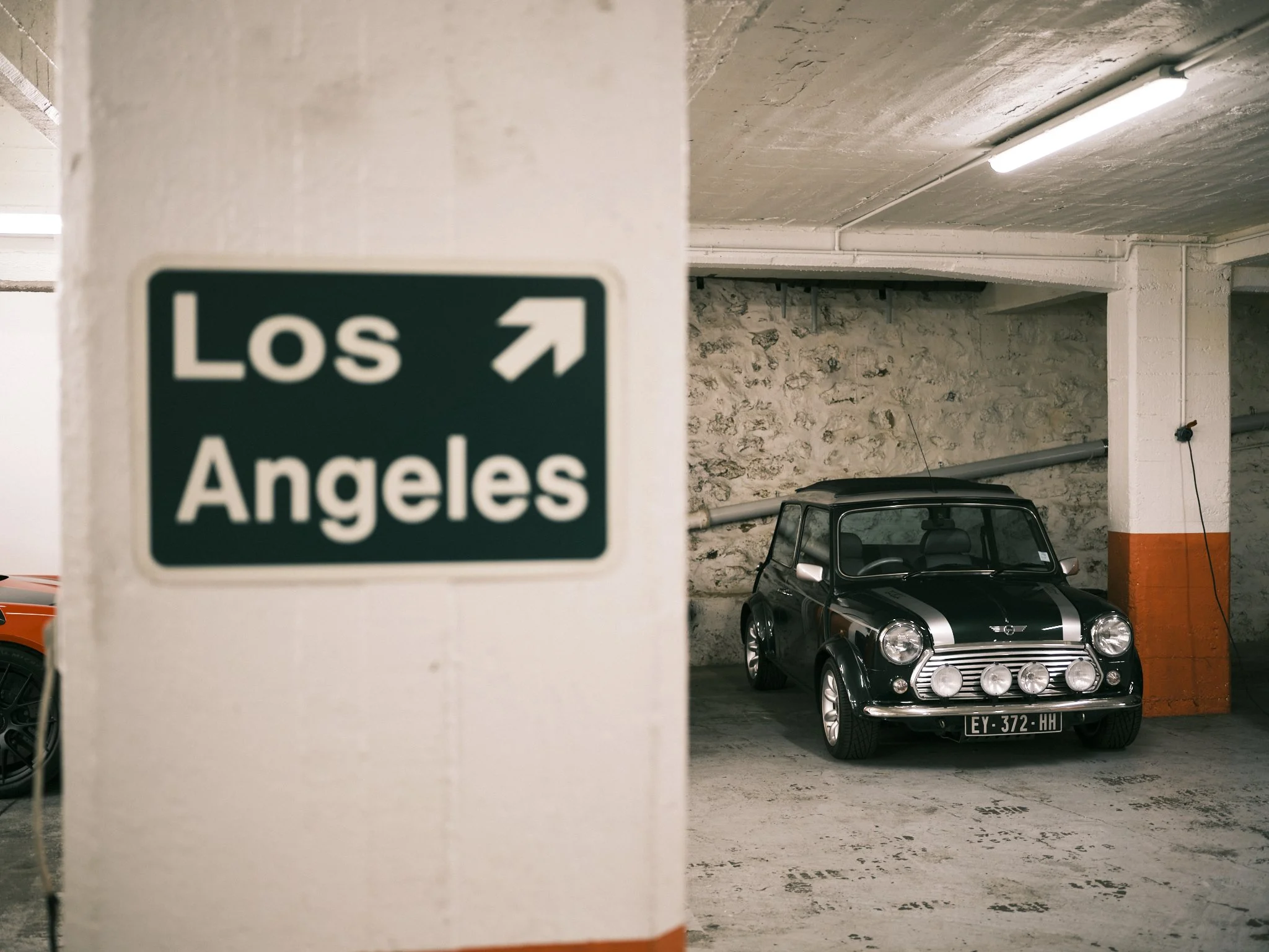 A parking garage with a small black car with white racing stripes parked near a stone wall, and a blue sign indicating directions to Los Angeles.
