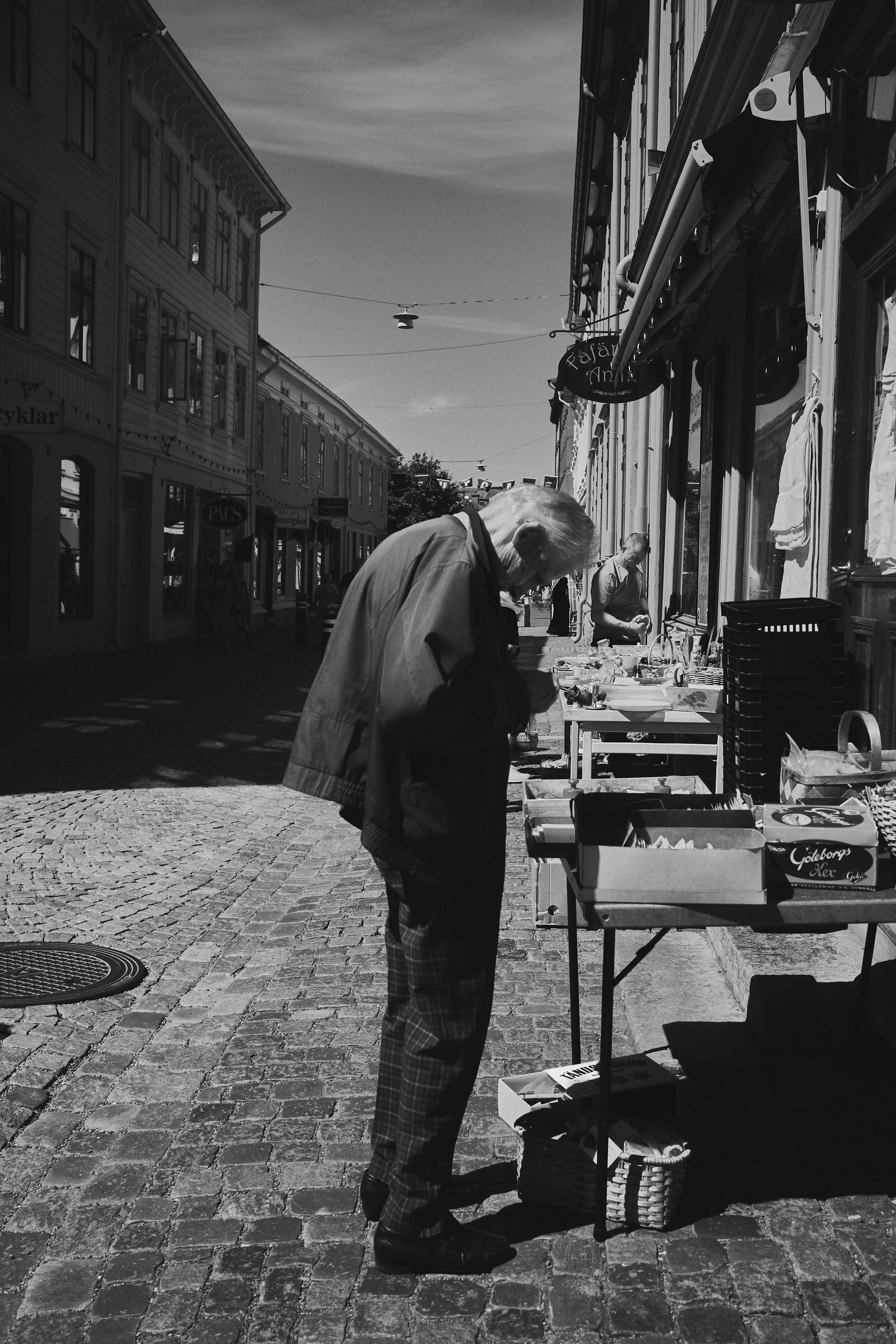 An elderly man browsing items at a street market stall on a cobblestone street in a European city, with other street vendors and pedestrians in the background.