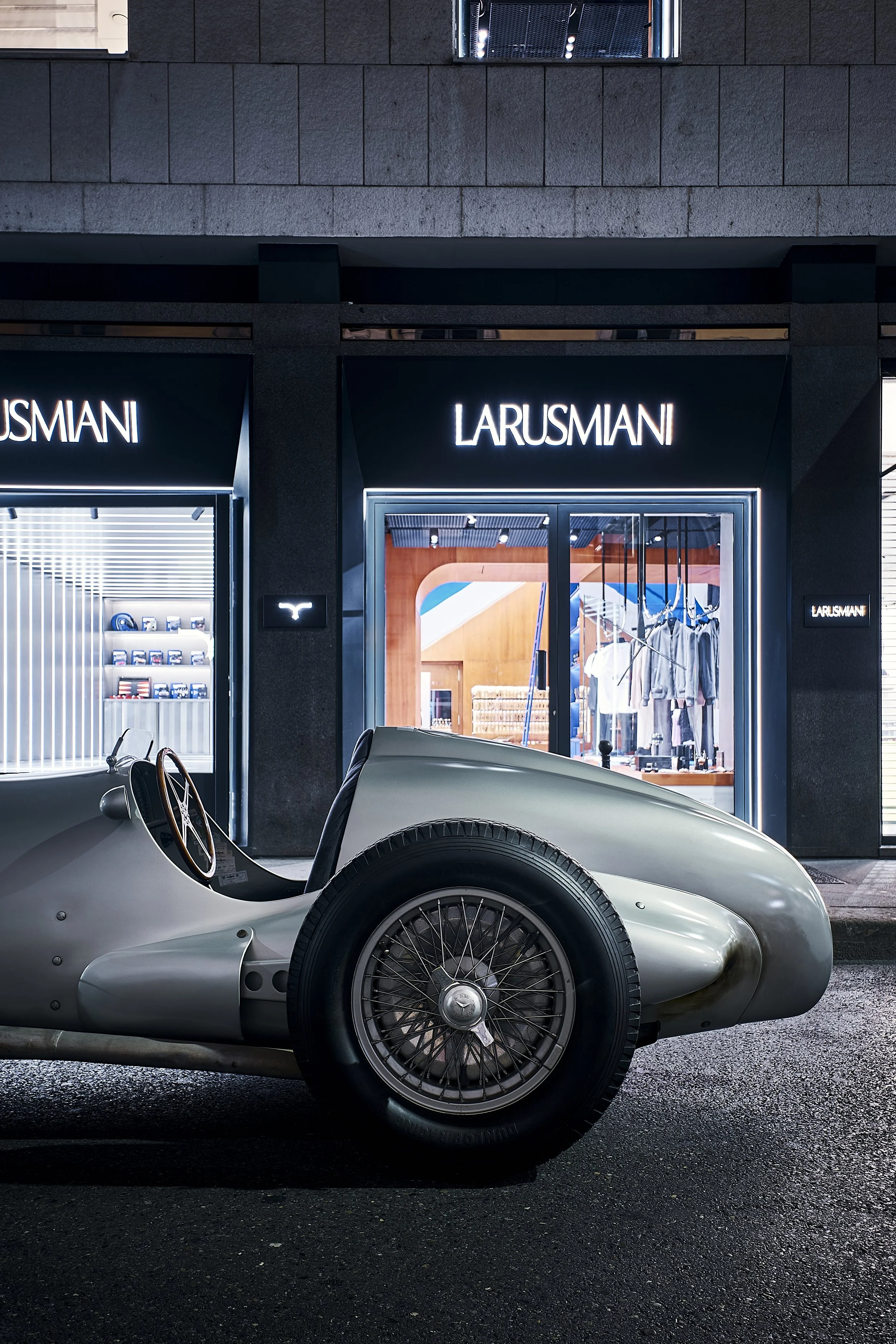 A vintage silver race car parked outside a La RUSMIANI store at night, with bright store lights illuminating the scene.