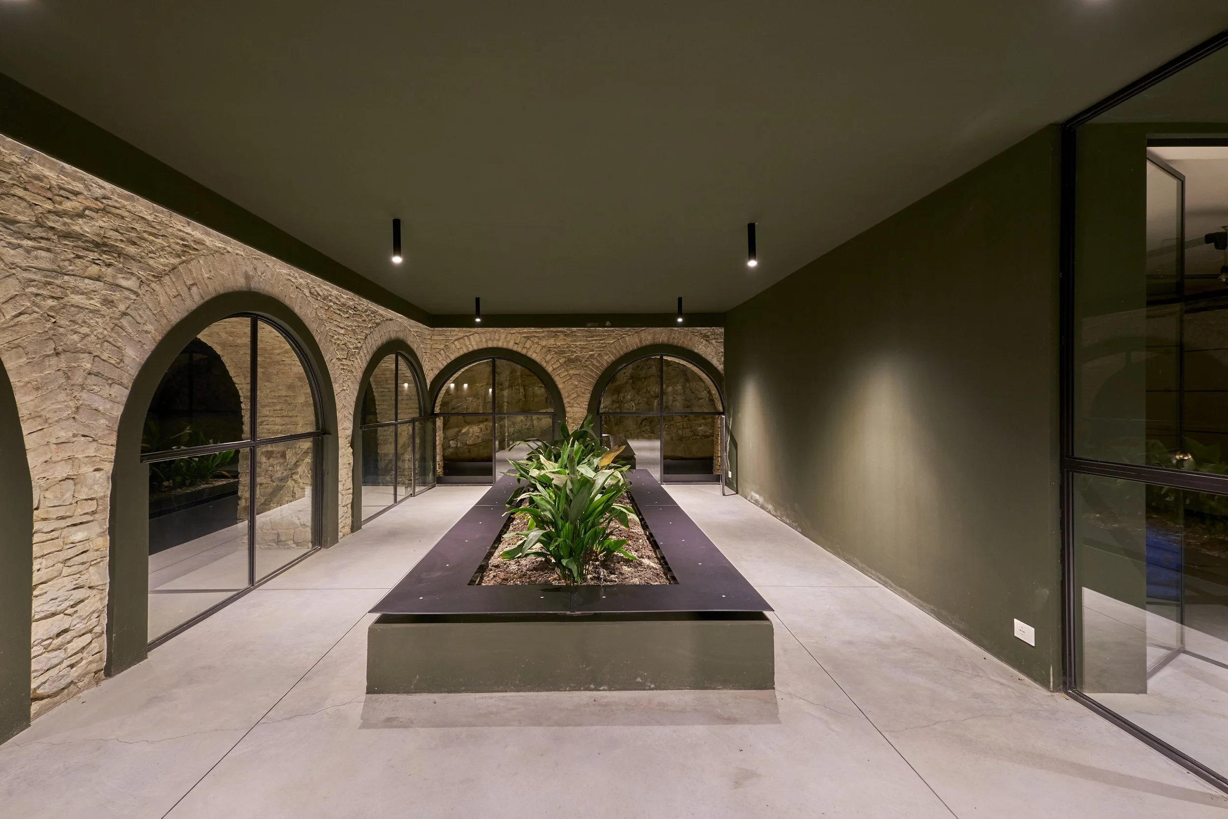 Modern indoor patio with stone wall arches, large glass windows, a rectangular planter with green plants, black ceiling lights, and concrete floors.