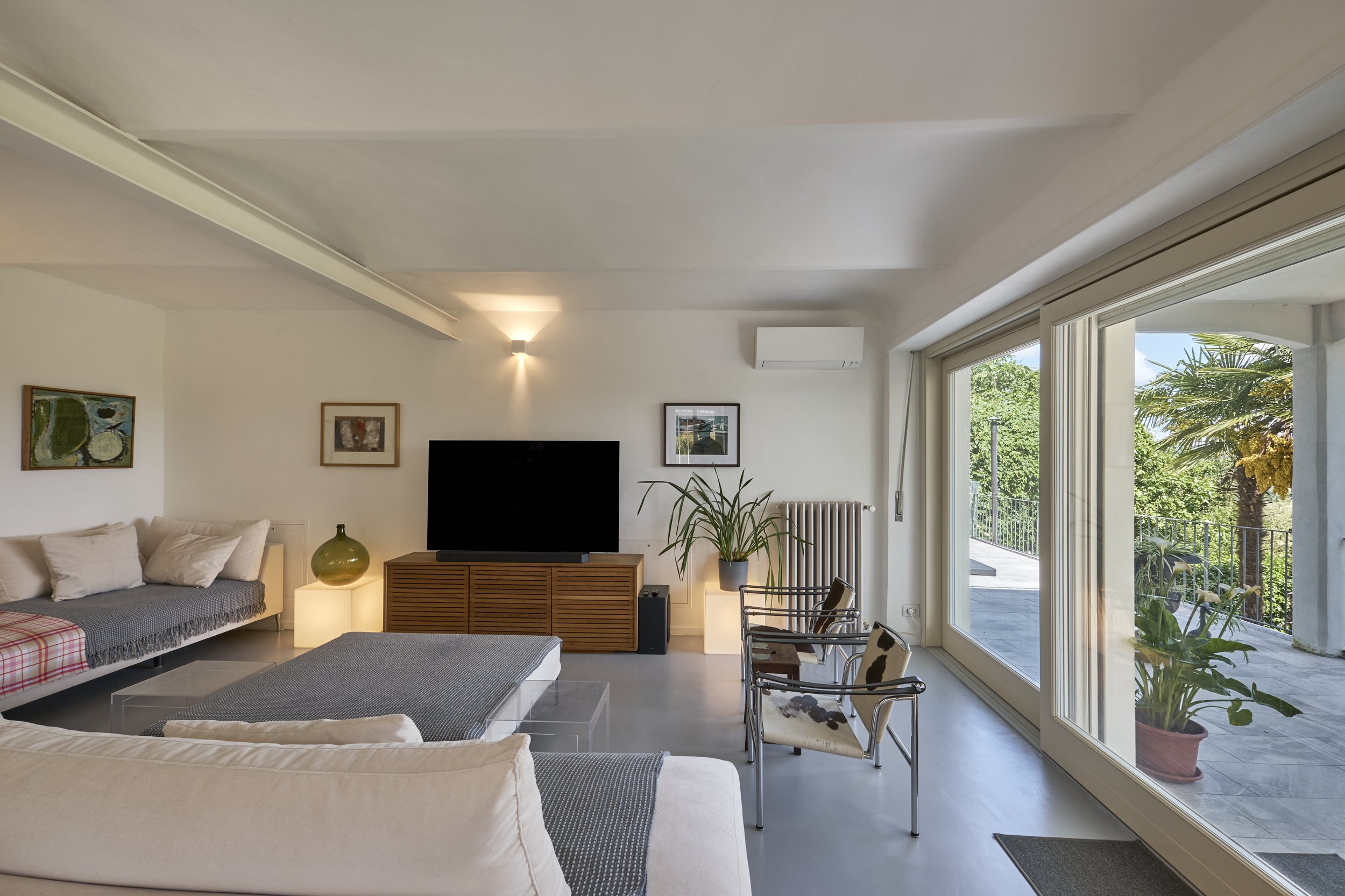 Living room with white walls, a large sliding glass door leading to an outdoor balcony, a white sofa, a coffee table, a television on a wooden console, two armchairs with cowhide cushions, potted plants, and framed artwork on the walls.