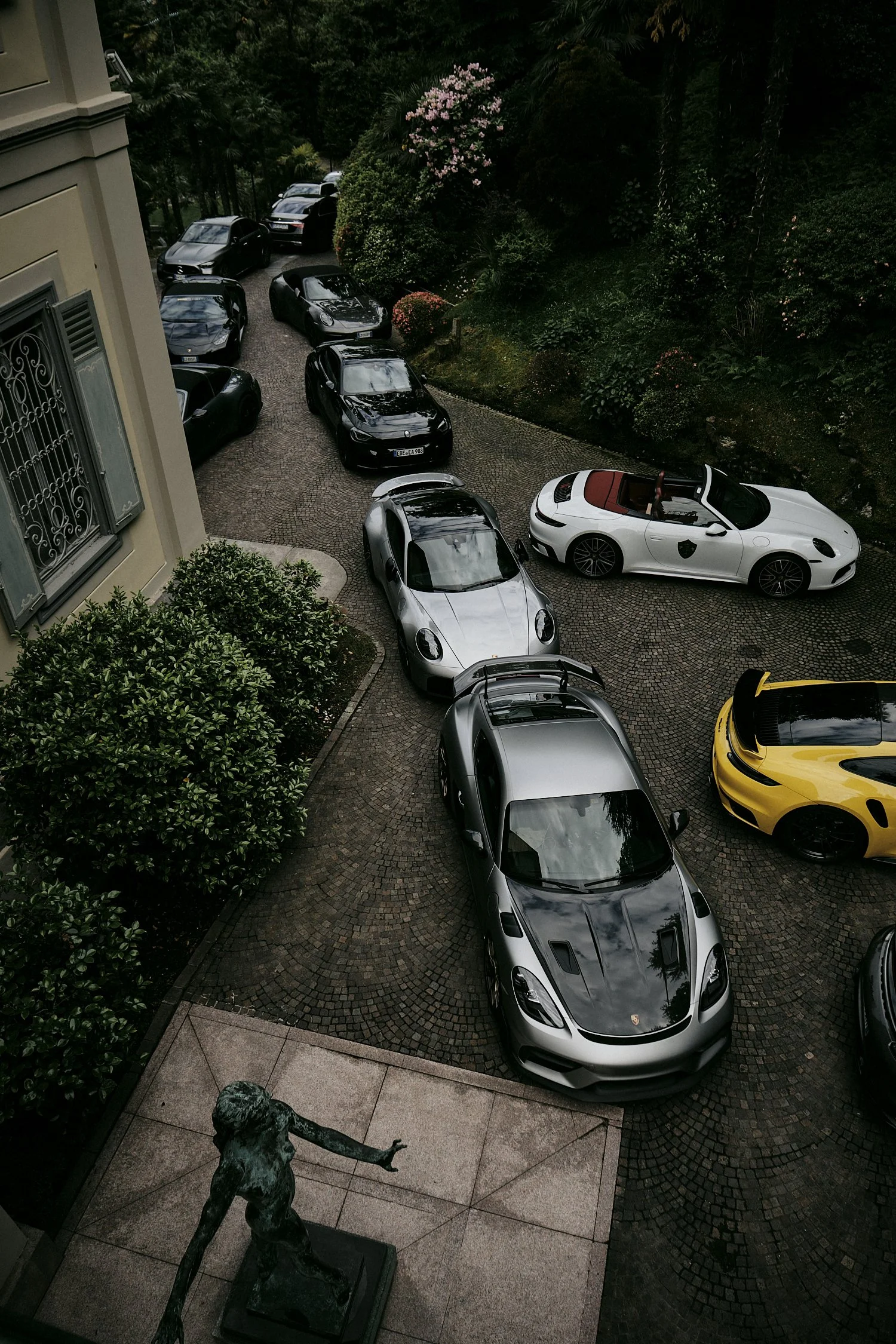 Multiple luxury cars parked on a cobblestone driveway next to a building with lush greenery and flowering bushes nearby.