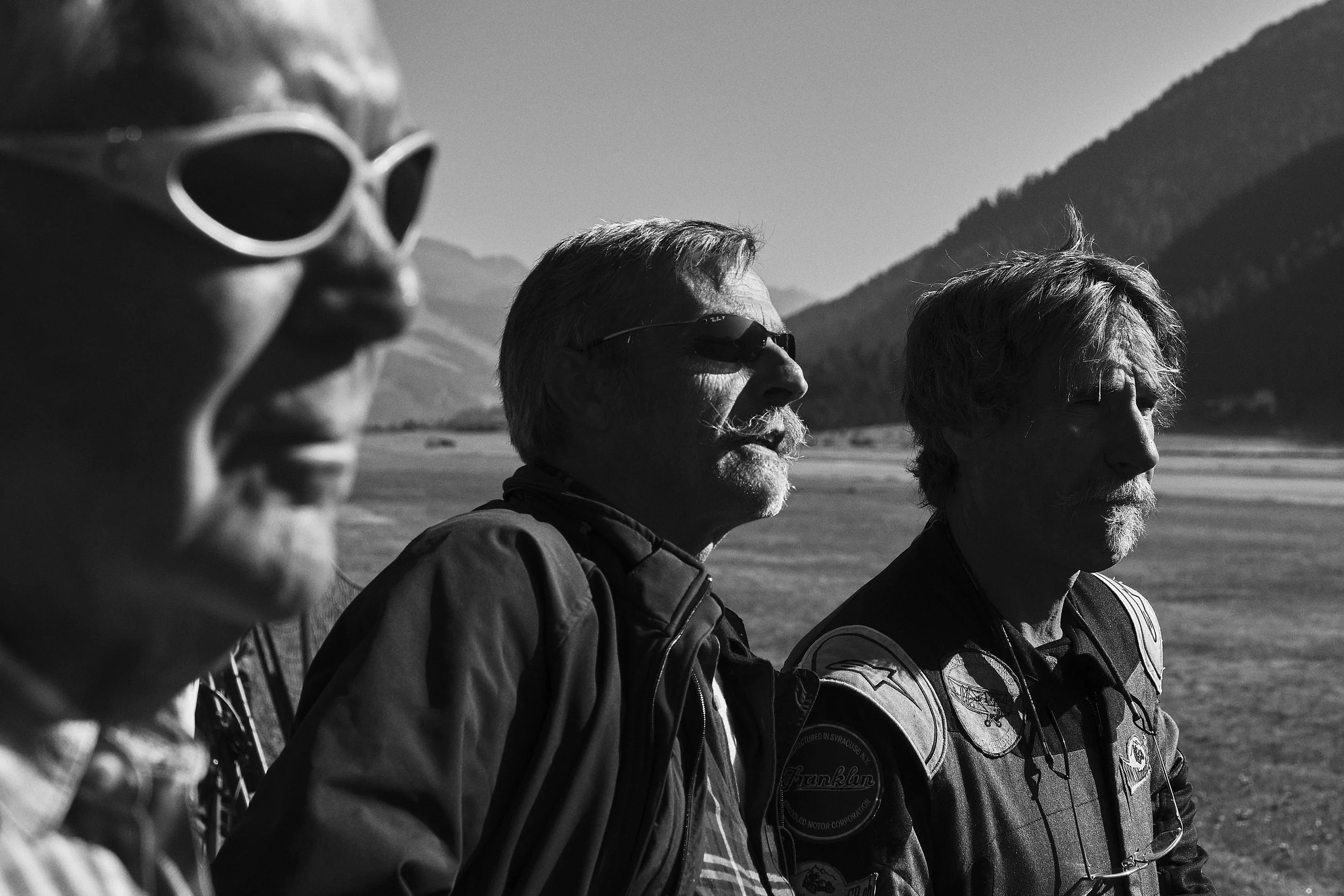 Three older individuals standing outdoors with mountains in the background, wearing sunglasses and motorcycle gear, possibly preparing for a ride.