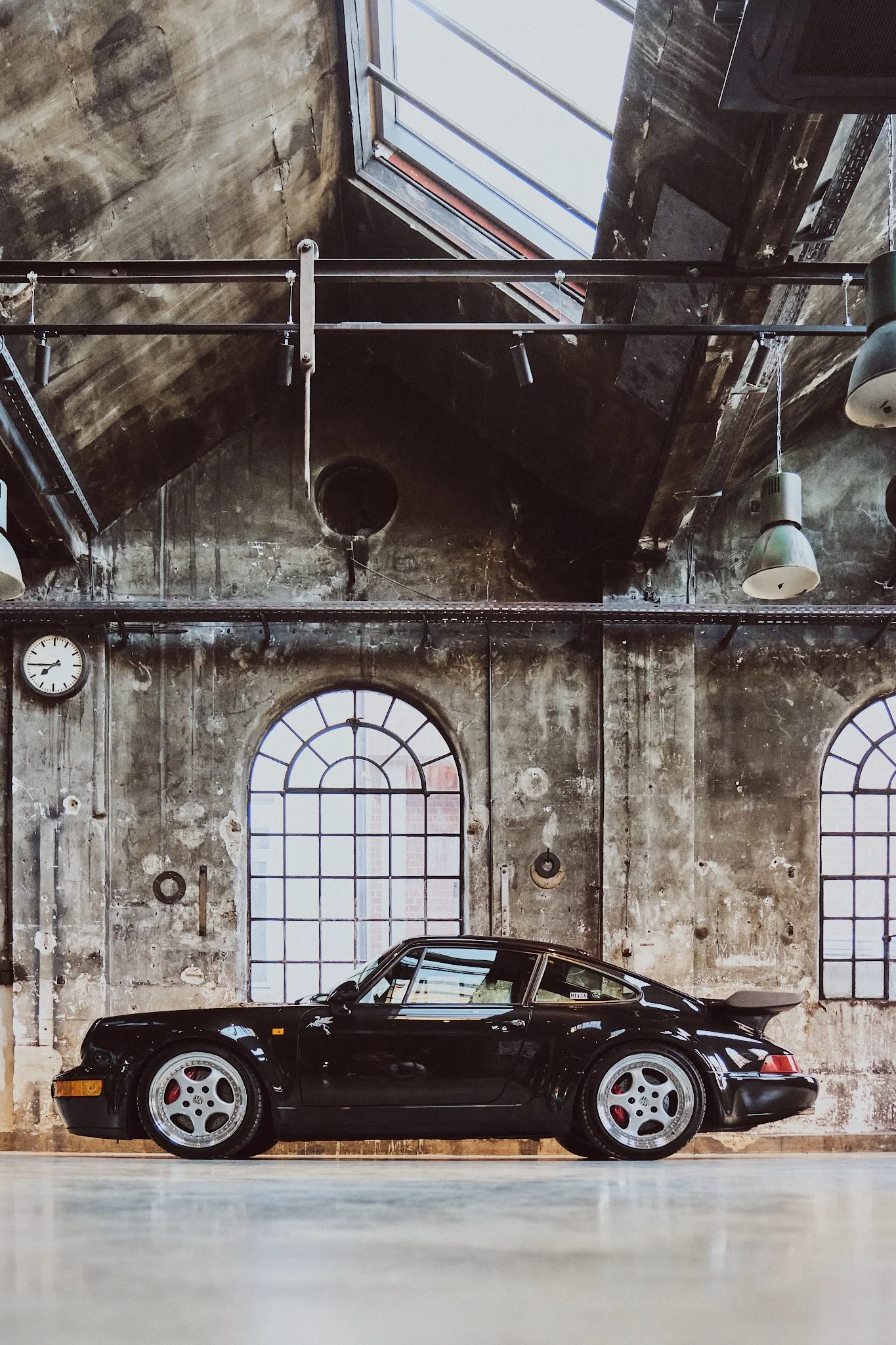 A black sports car parked indoors against a weathered industrial-style brick wall with large arched windows and an exposed ceiling with lighting fixtures.