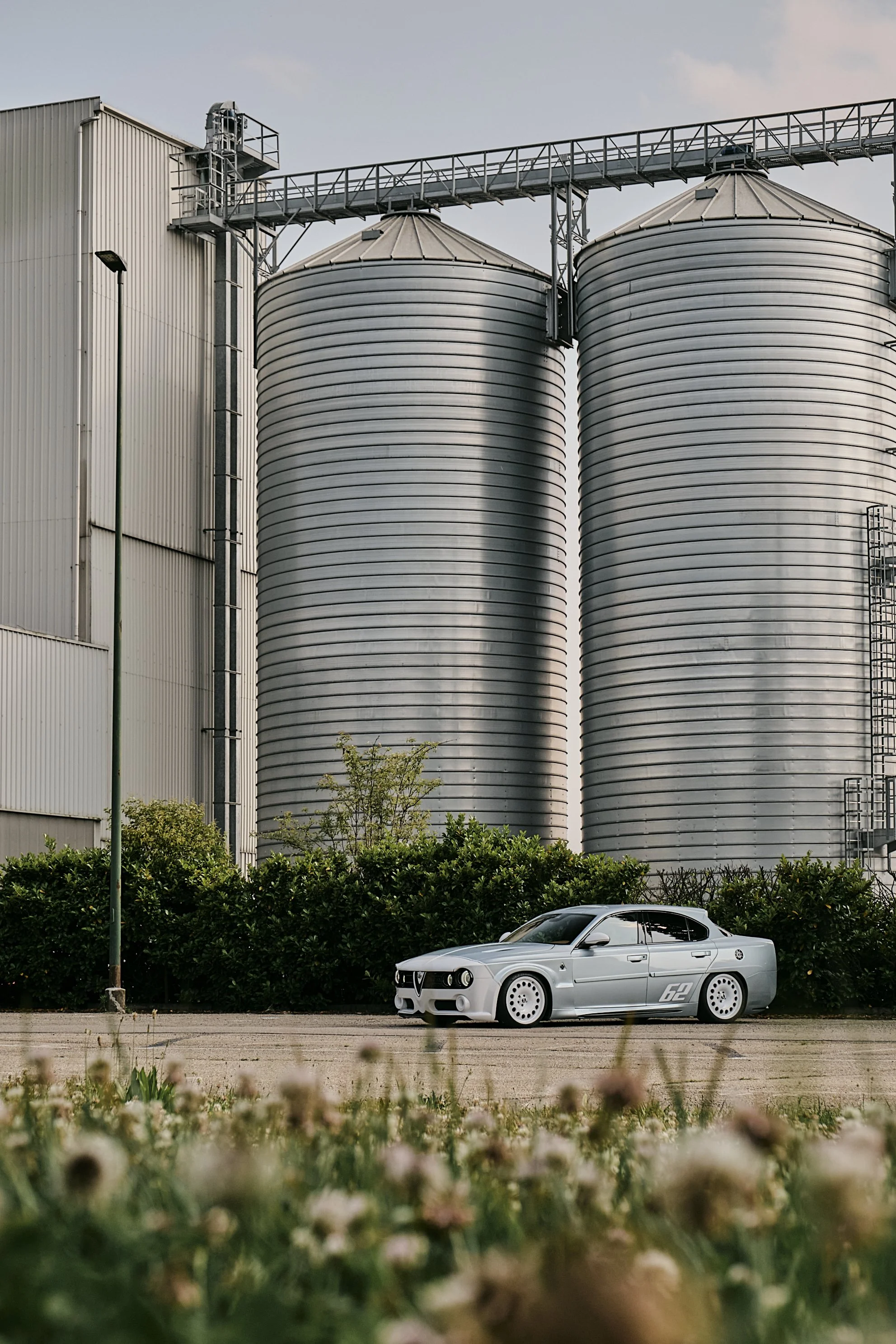 A silver sports car parked near large industrial silos with green bushes and a flowered foreground.