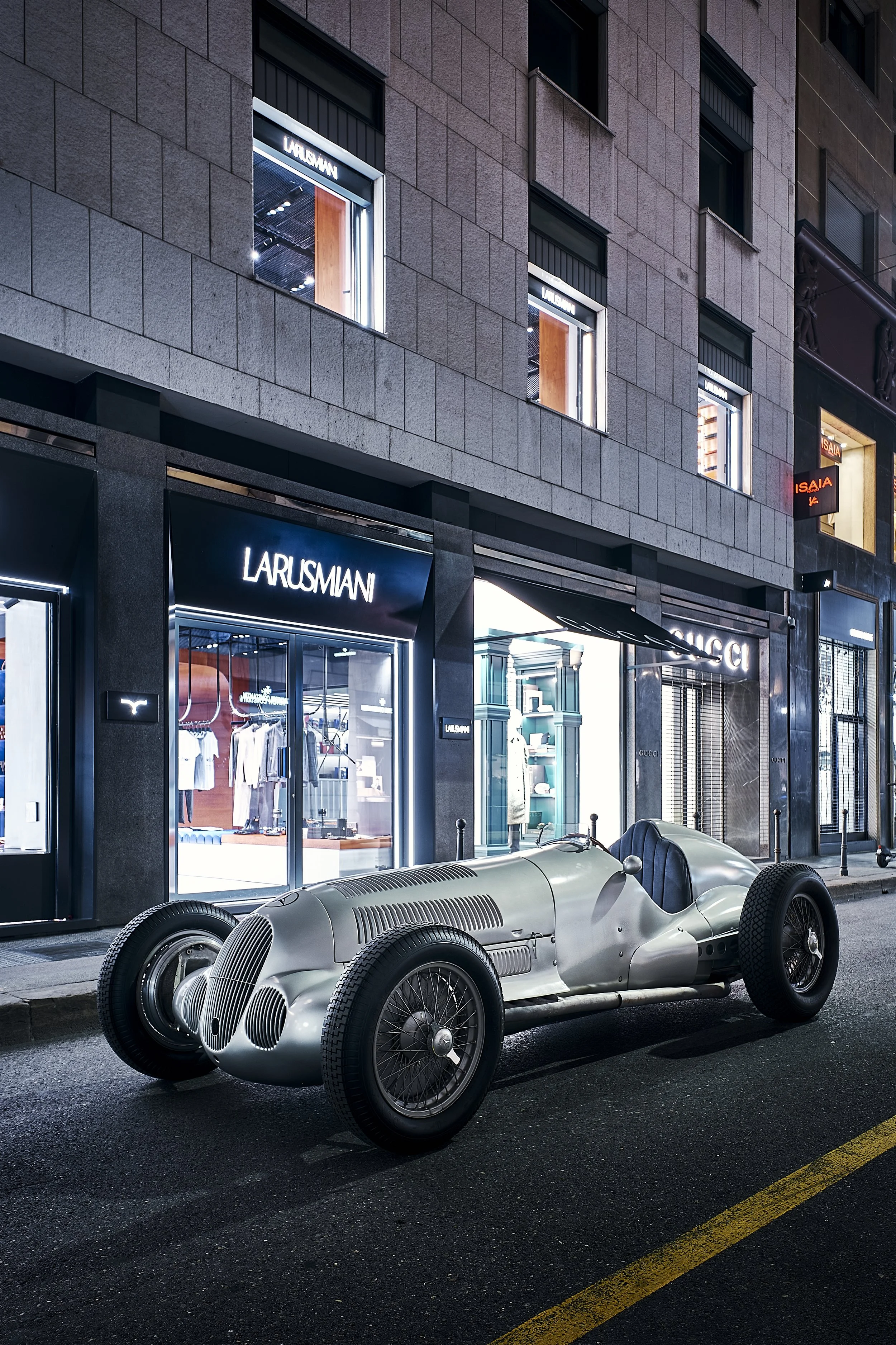 Vintage silver racing car parked in front of high-end fashion stores, including Larusmiani and Gucci, on a city street at night.