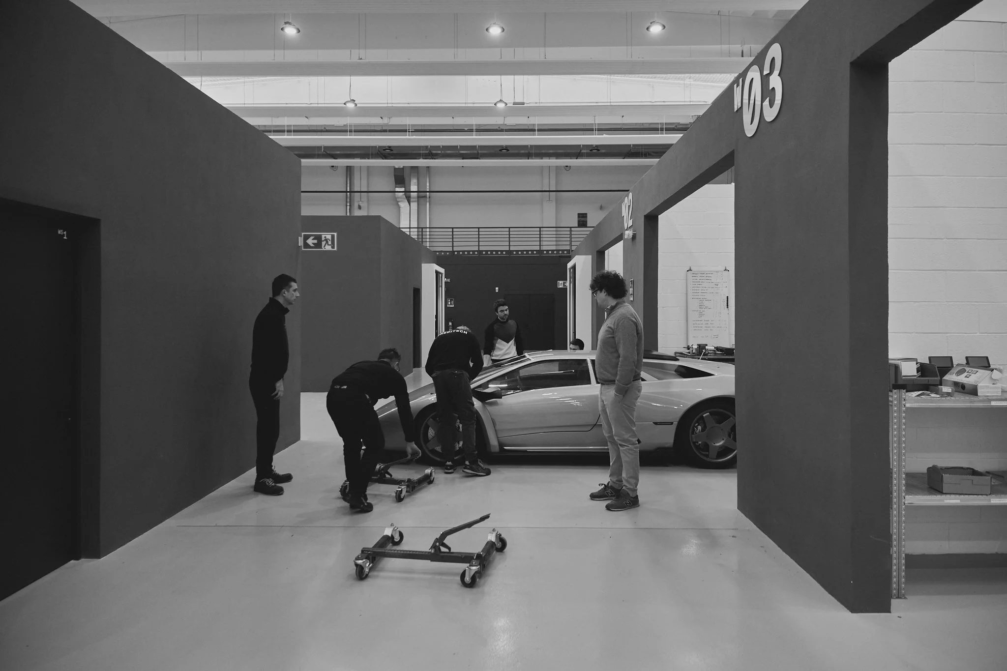 Black and white photo of a modern car showroom with a sports car being assembled or prepared. Five people are present, some working on or inspecting the car, which is positioned between partition walls labeled 02 and 03. Tools and equipment are visib