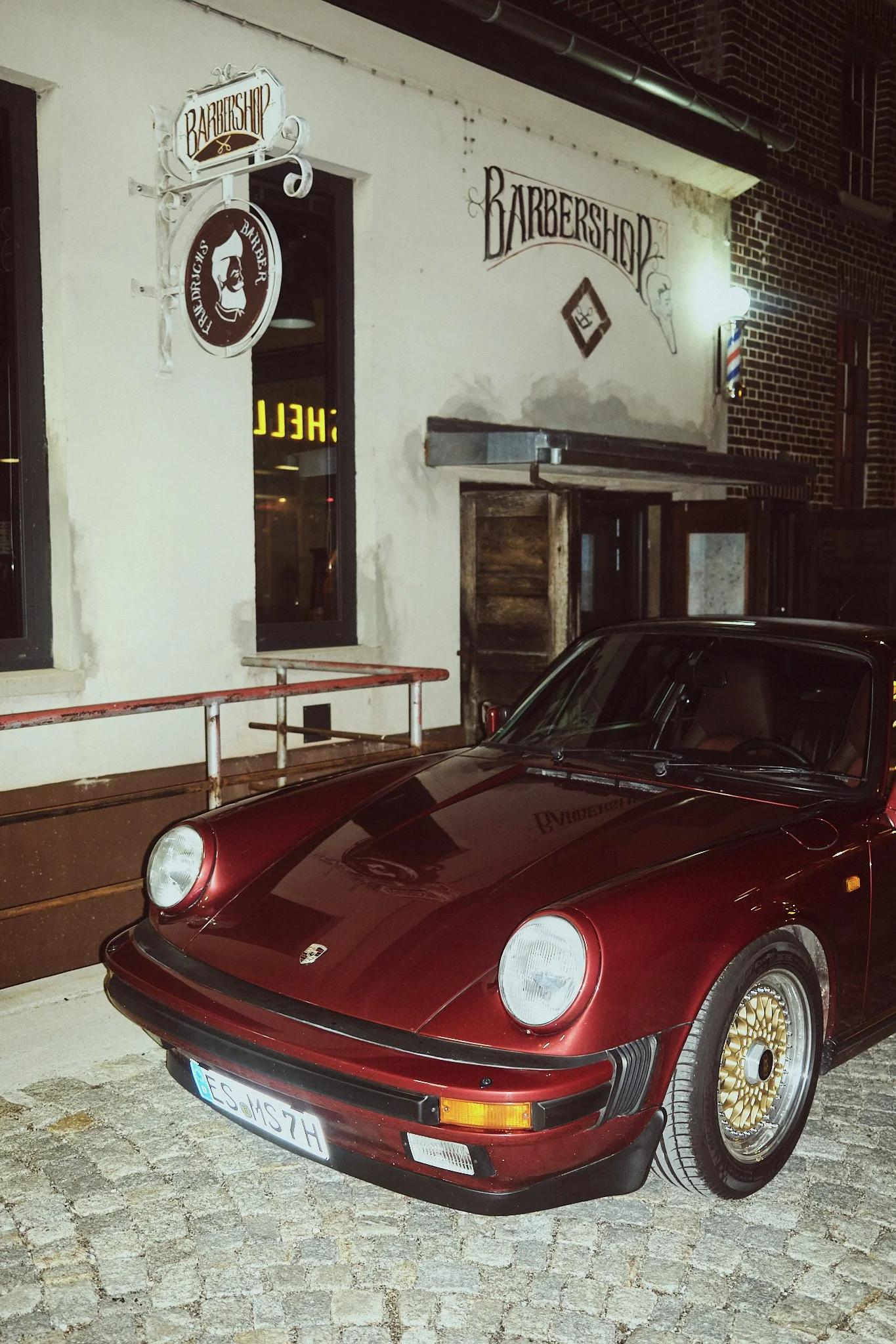 A vintage red Porsche parked outside a barber shop at night. The shop has signs on the wall that read 'Barbershop', with a barber pole near the entrance.