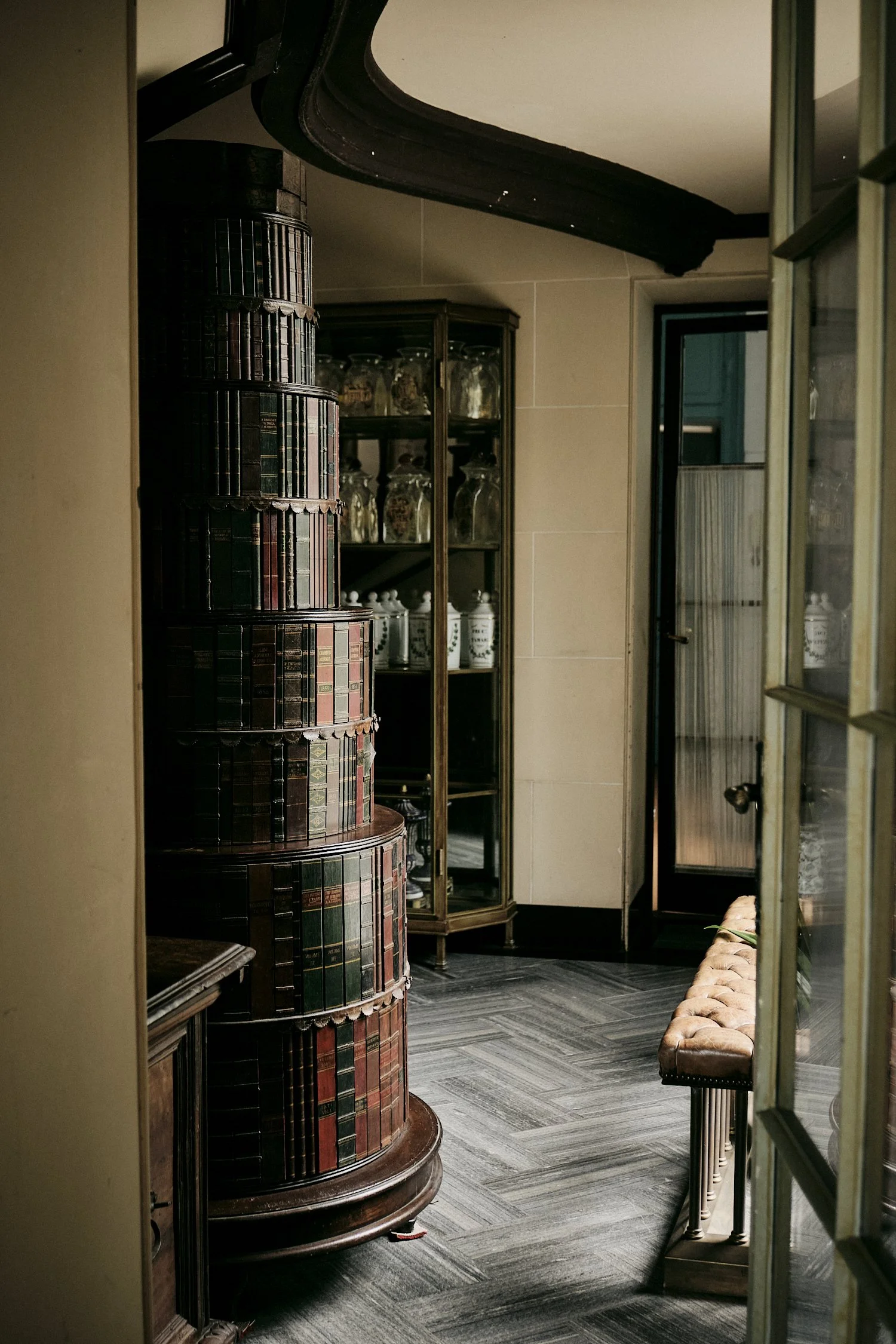 Interior of a vintage room with dark wood furniture, a curved cabinet decorated with books, a glass display cabinet, a bench, and a door leading to a balcony, with sunlight illuminating the space.