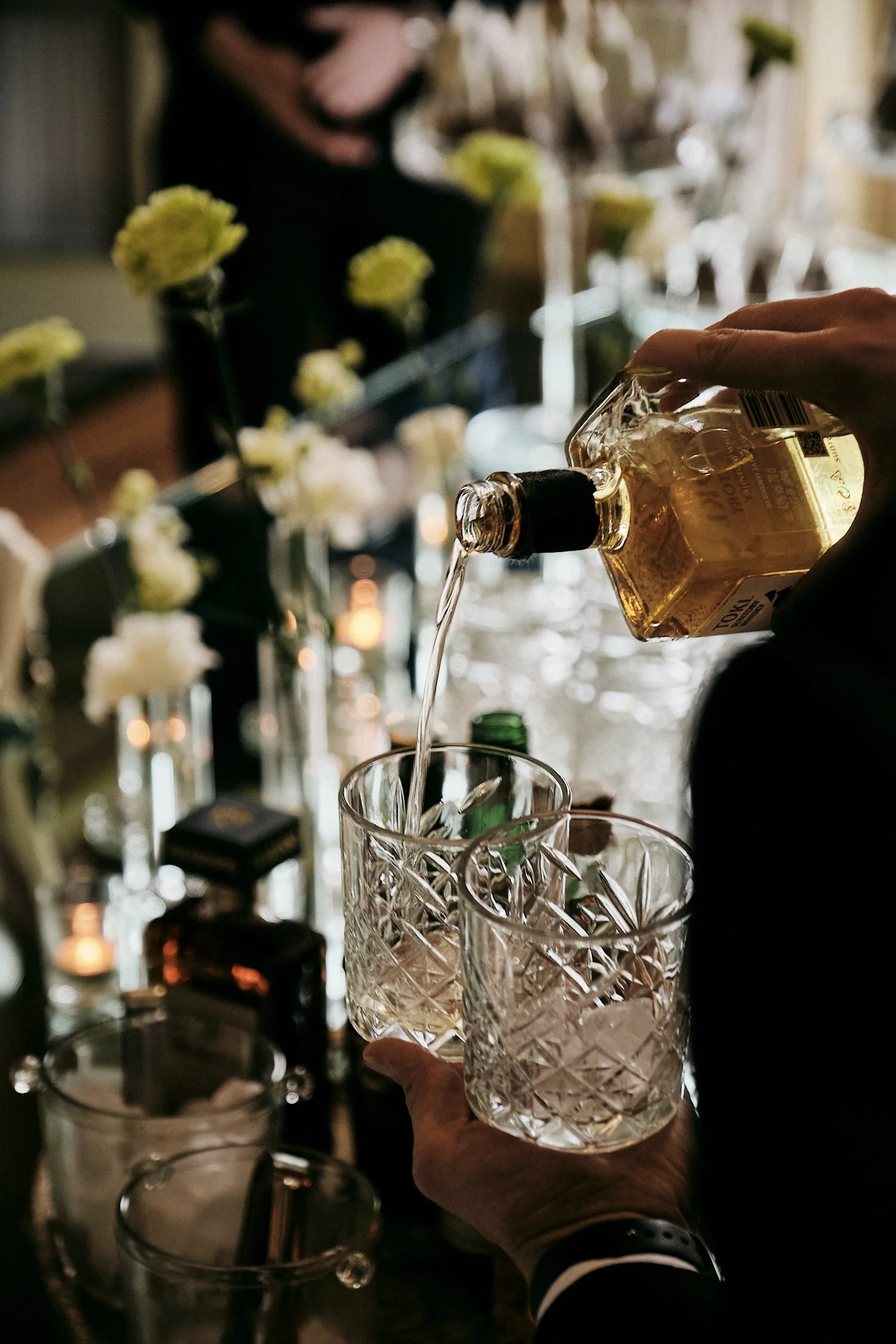 Person pours whiskey into crystal glass at a bar with floral decorations in the background.