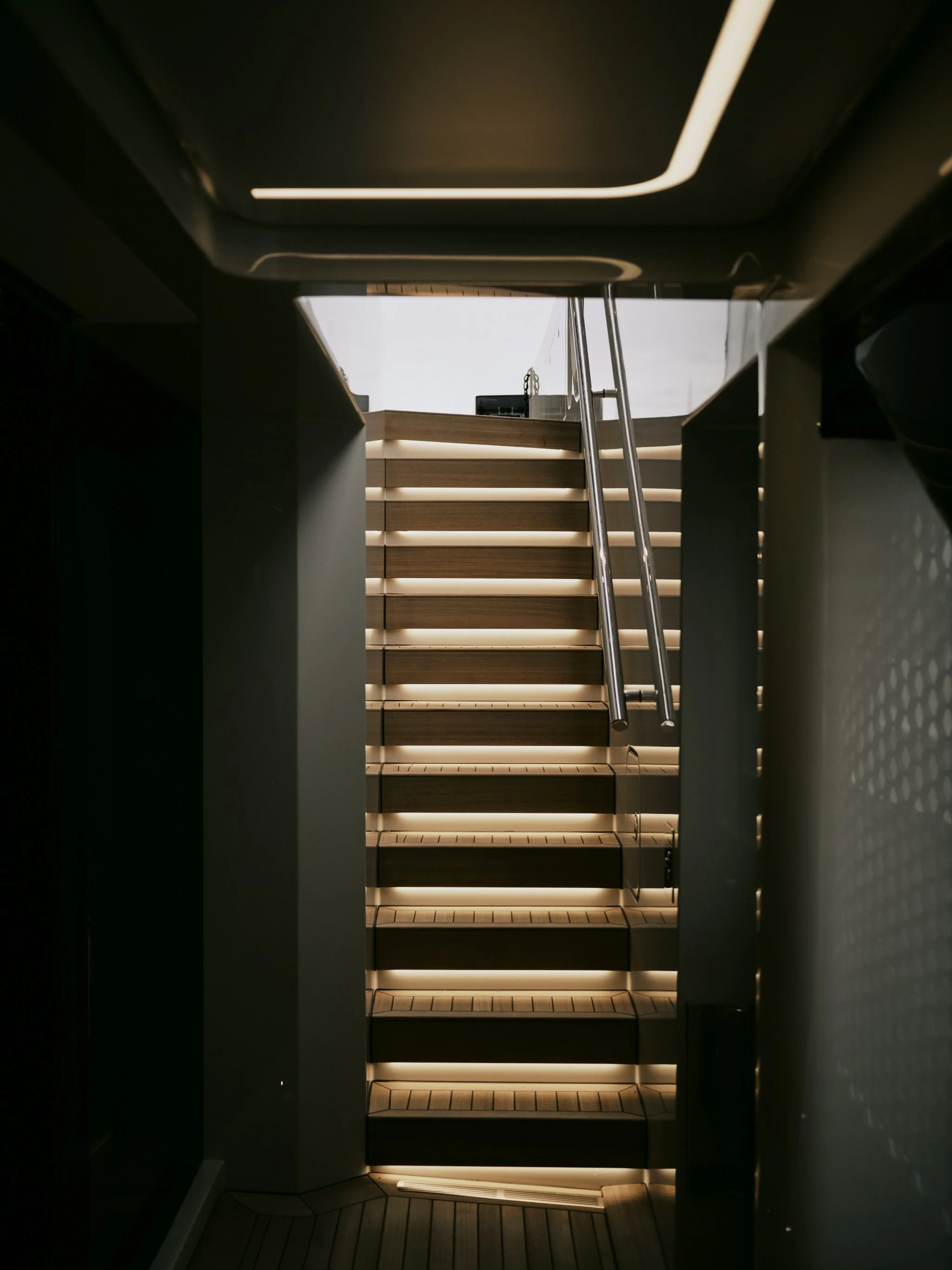 Wooden staircase with integrated lighting and metallic handrails leading to an upper level, viewed from a dark hallway.