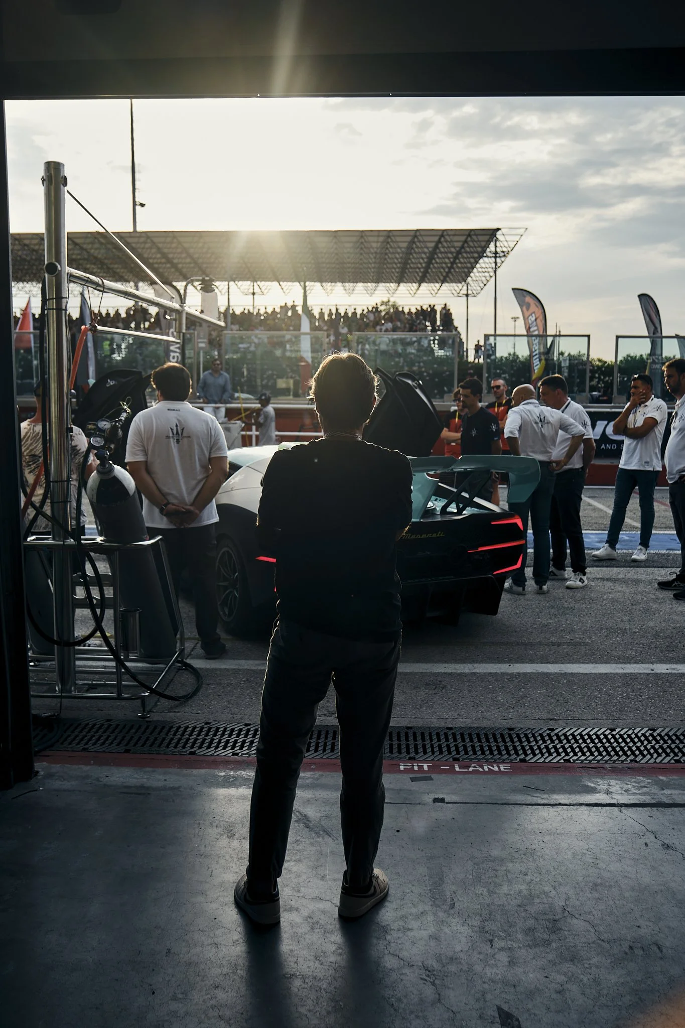 Person standing in a garage, observing a race car being prepared in the pit lane at a motorsport event with spectators and team members in the background.