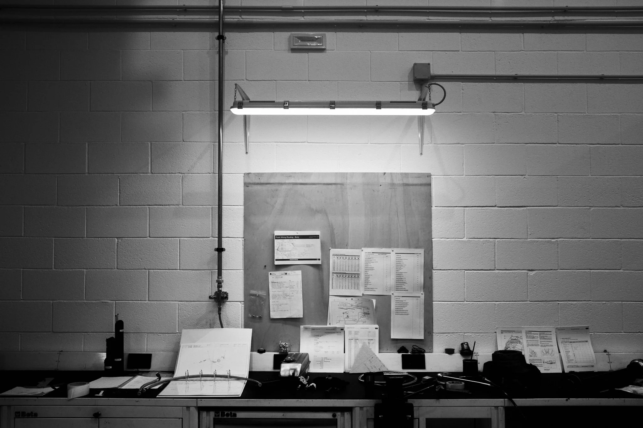 A black-and-white photo of a workbench against a cinderblock wall with various papers, tools, and equipment, illuminated by a single fluorescent light fixture.