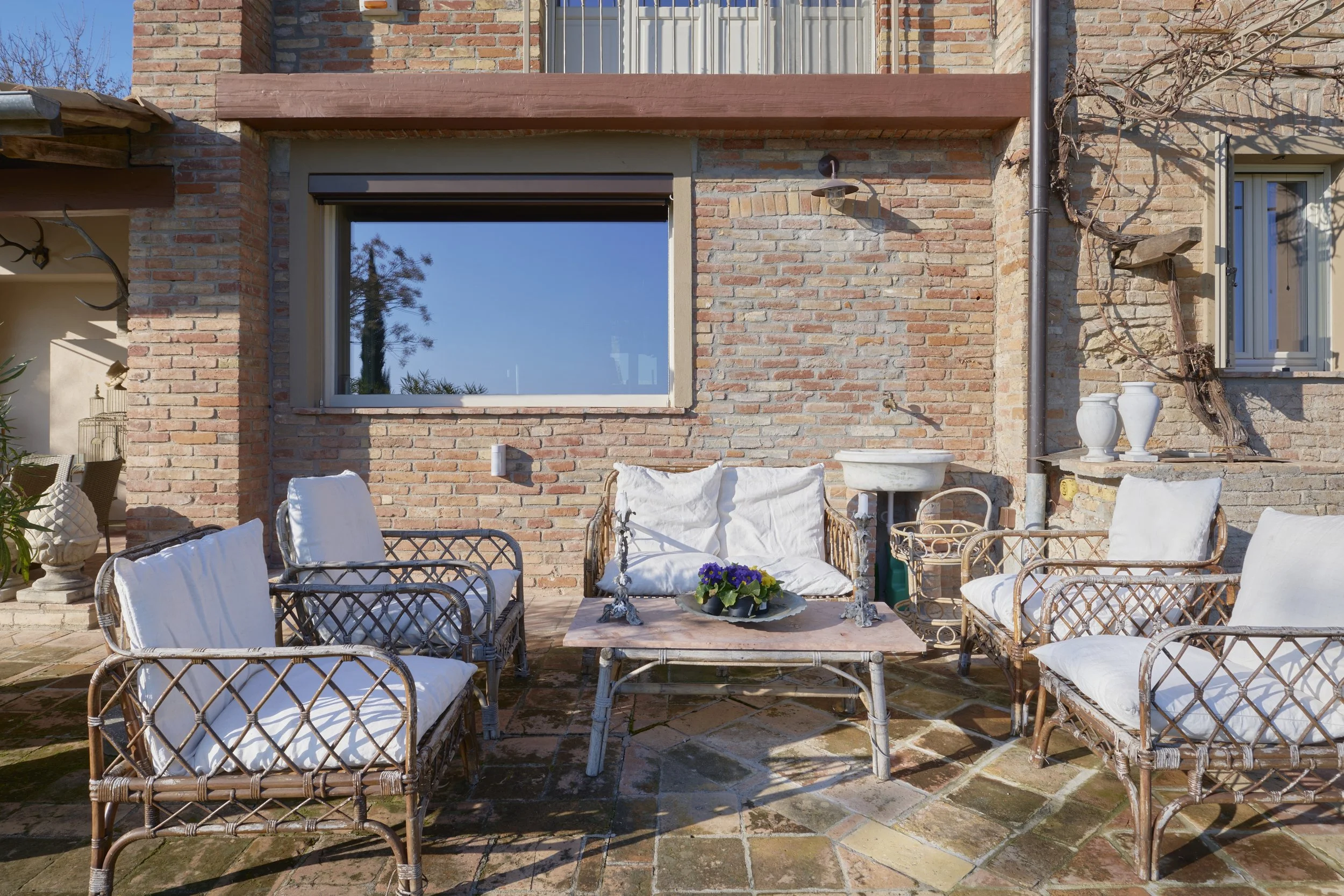 Outdoor patio with wicker furniture including chairs with white cushions, a table with a flower arrangement, against a brick wall with windows and decorative vases.