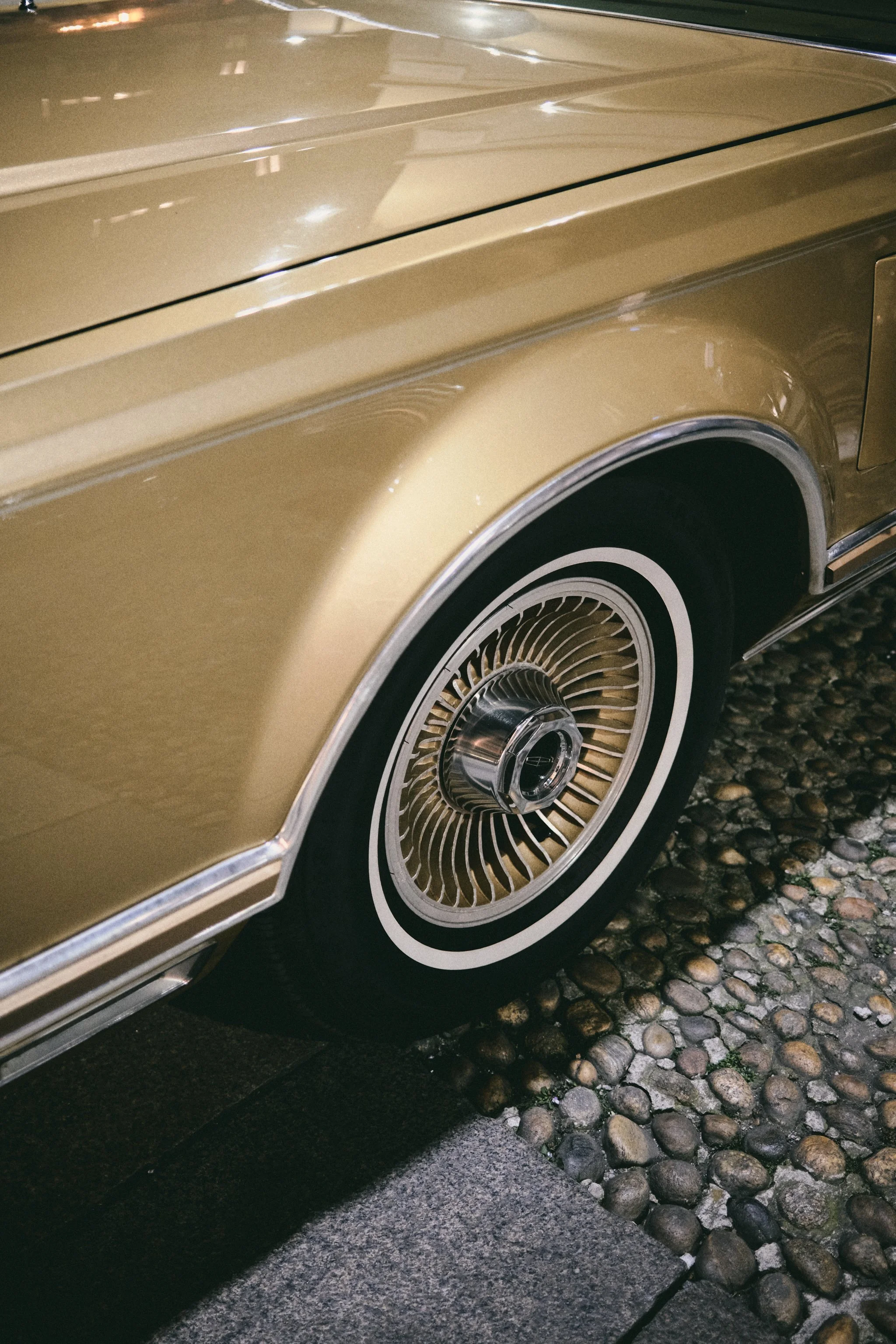 Close-up of a vintage gold-colored car wheel with a wire-spoke rim and a black tire, parked on a cobblestone surface.