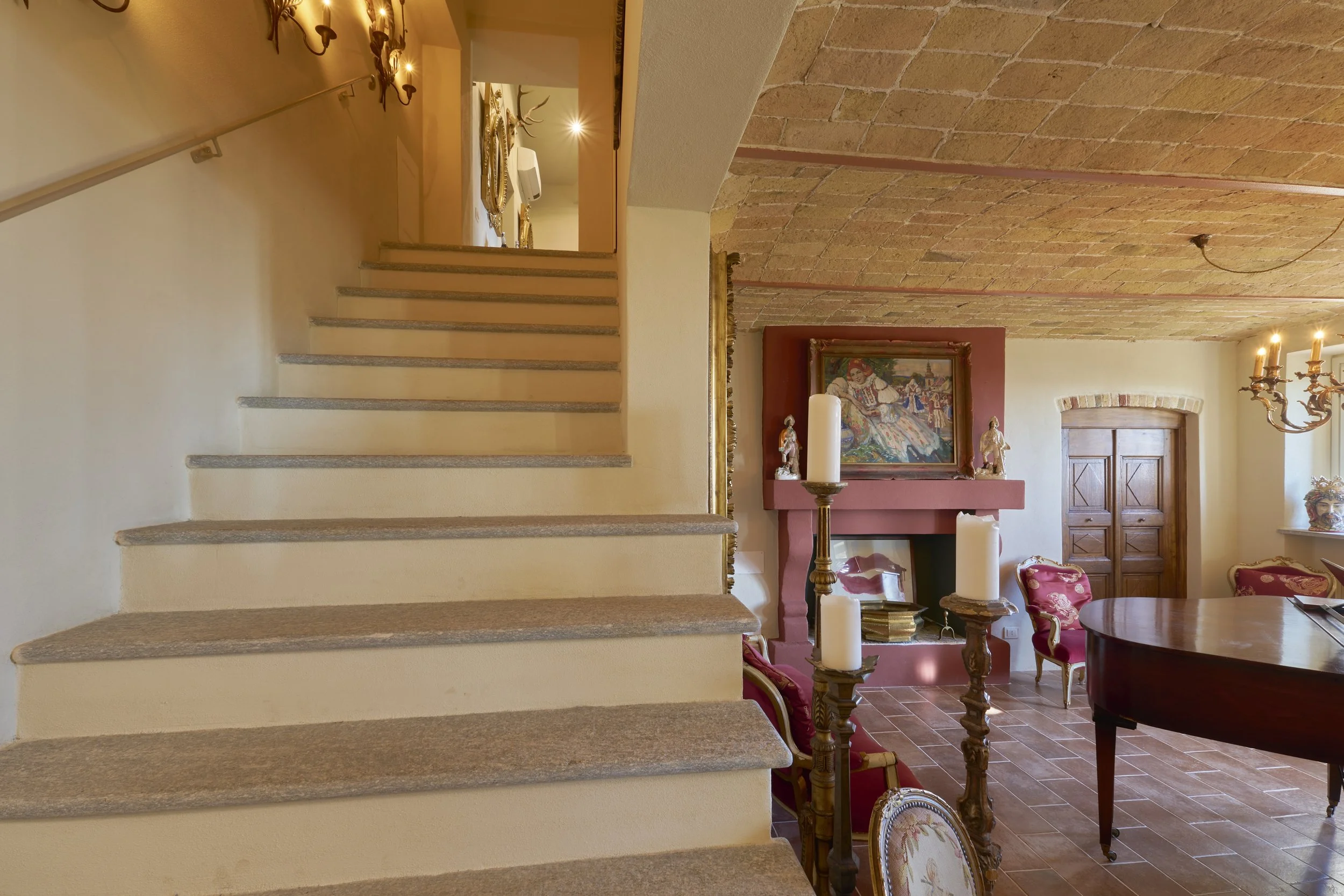 Interior of a room with a staircase on the left, a brick ceiling, a fireplace with artwork above, and elegant furniture including chairs, a piano, and candles.