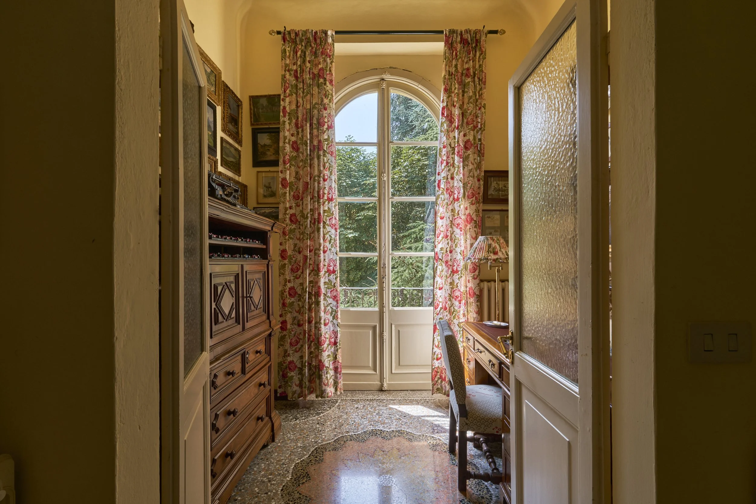 A home office with a large arched window covered by floral curtains, wooden furniture including a desk and cabinet, a chair, a table lamp, and several framed pictures on the wall, with sunlight streaming in.