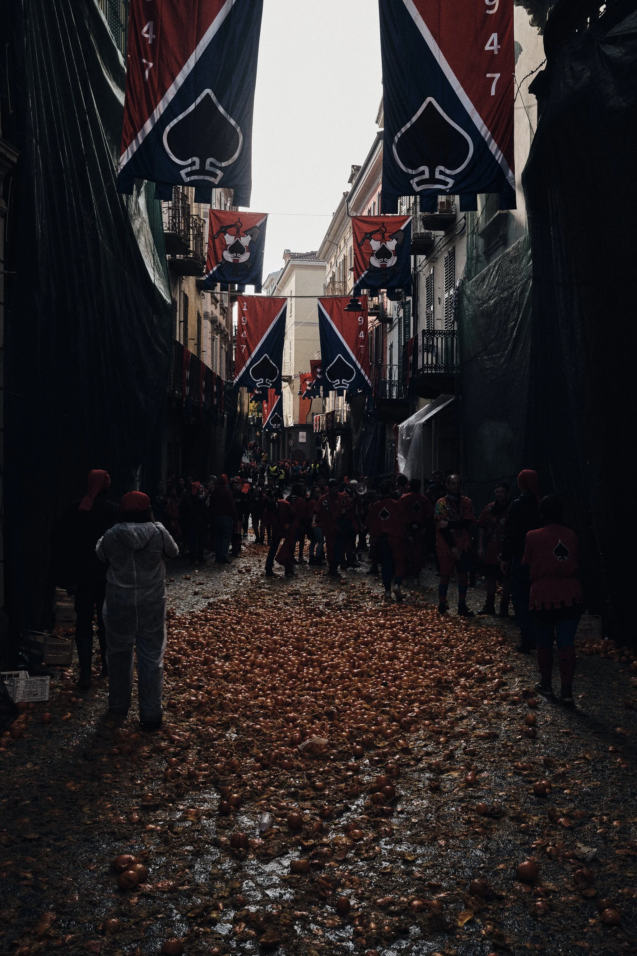 A street decorated with red, white, and blue banners featuring a spade symbol, with people wearing red and black clothing, some with spade symbols, amidst a messy ground covered with onions.