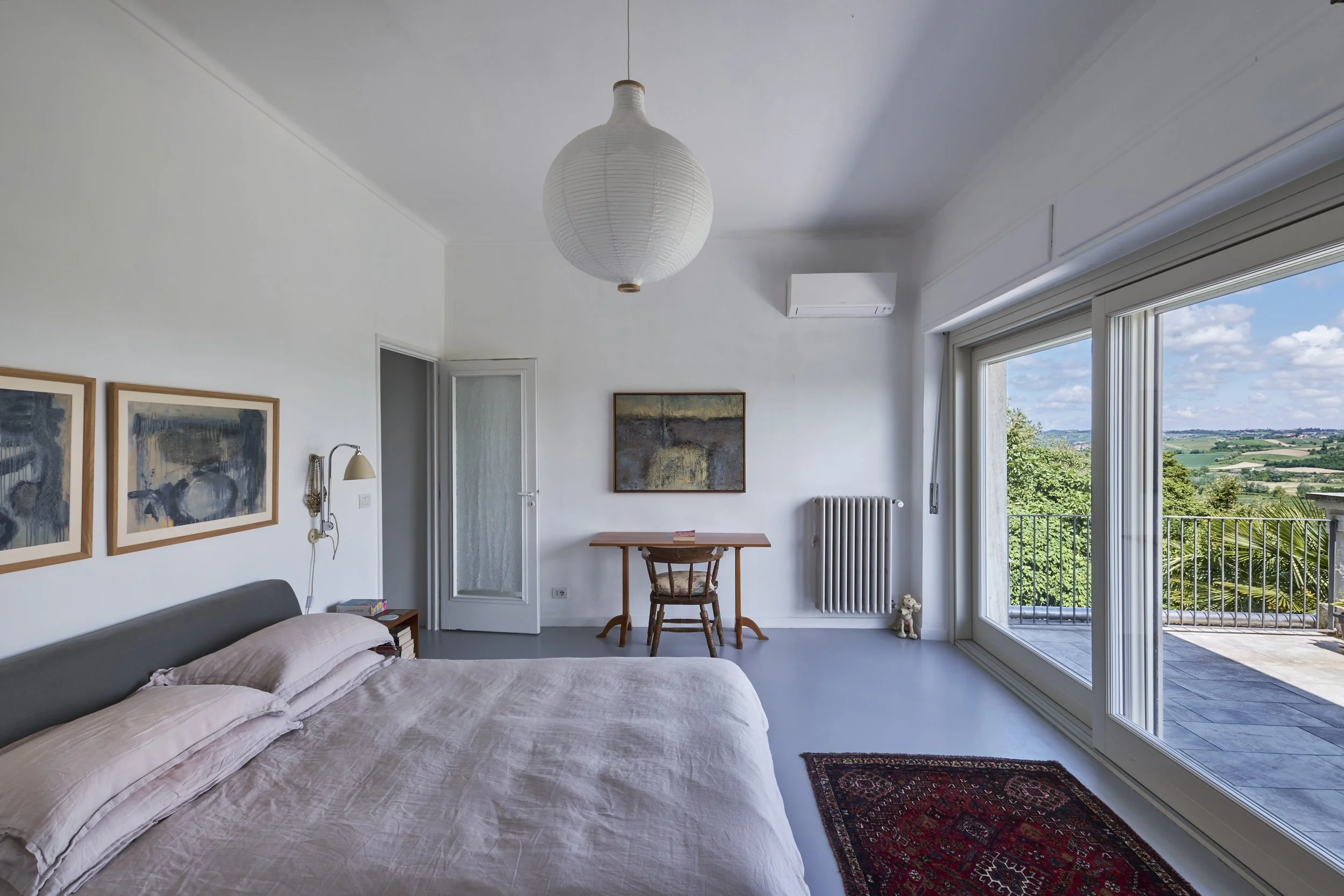 Bright bedroom with large sliding glass door overlooking a scenic landscape, bed with pinkish linens, artwork on white walls, wooden desk with chair, air conditioning unit, and a patterned rug on the floor.