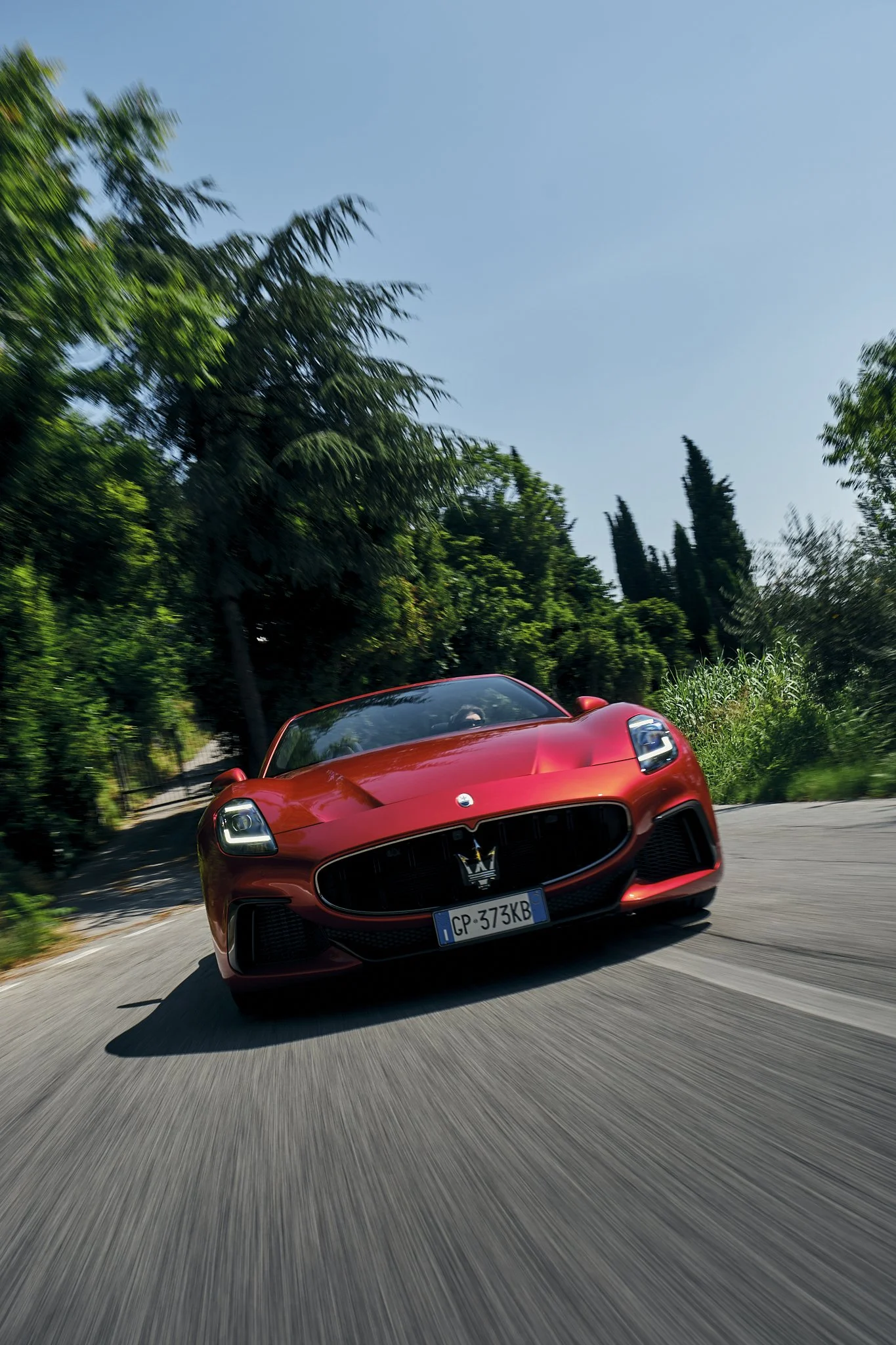 A red Maserati sports car driving on a winding road with green trees and blue sky in the background.