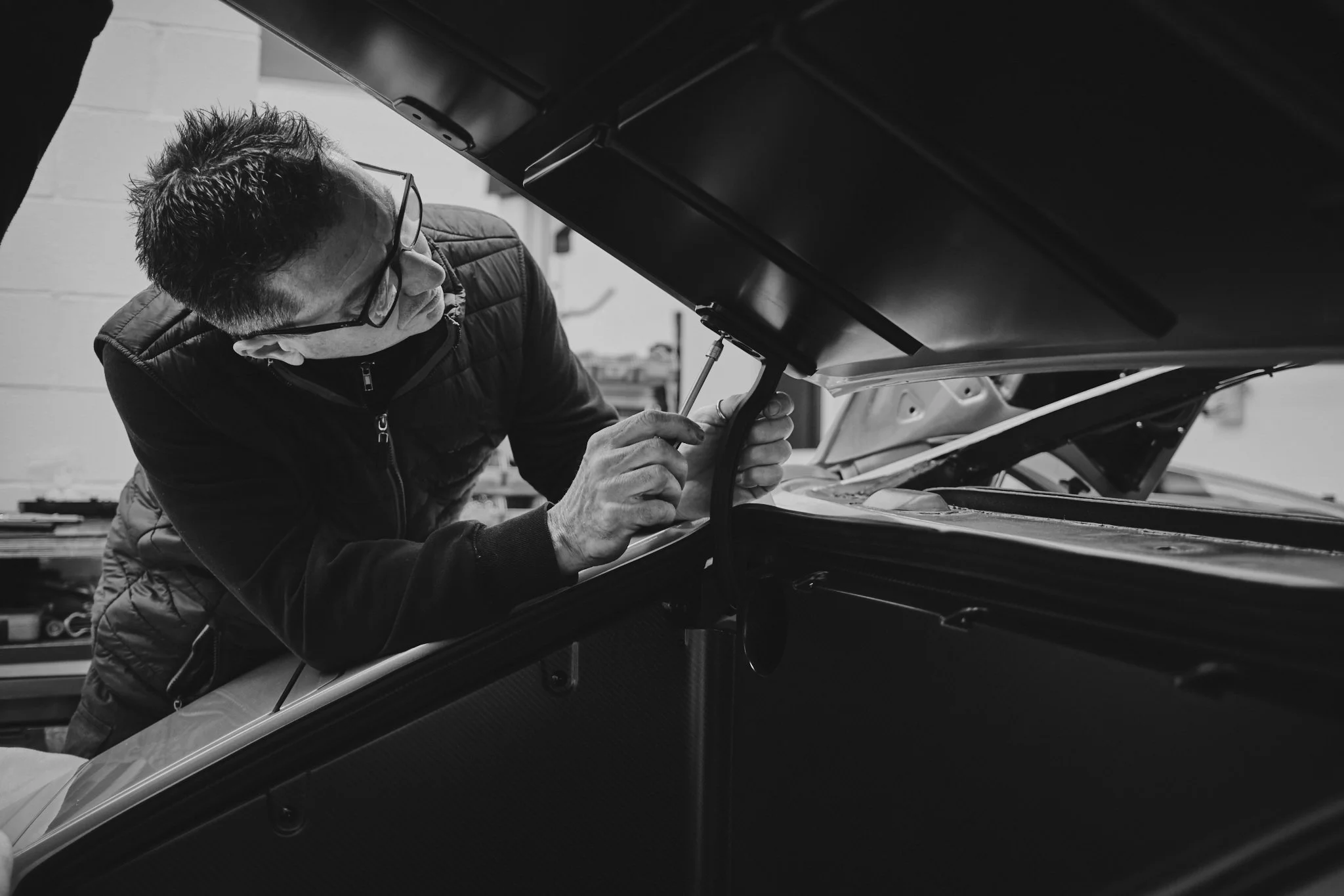A man working on a car engine with the hood open, wearing glasses and a face mask, in a workshop.