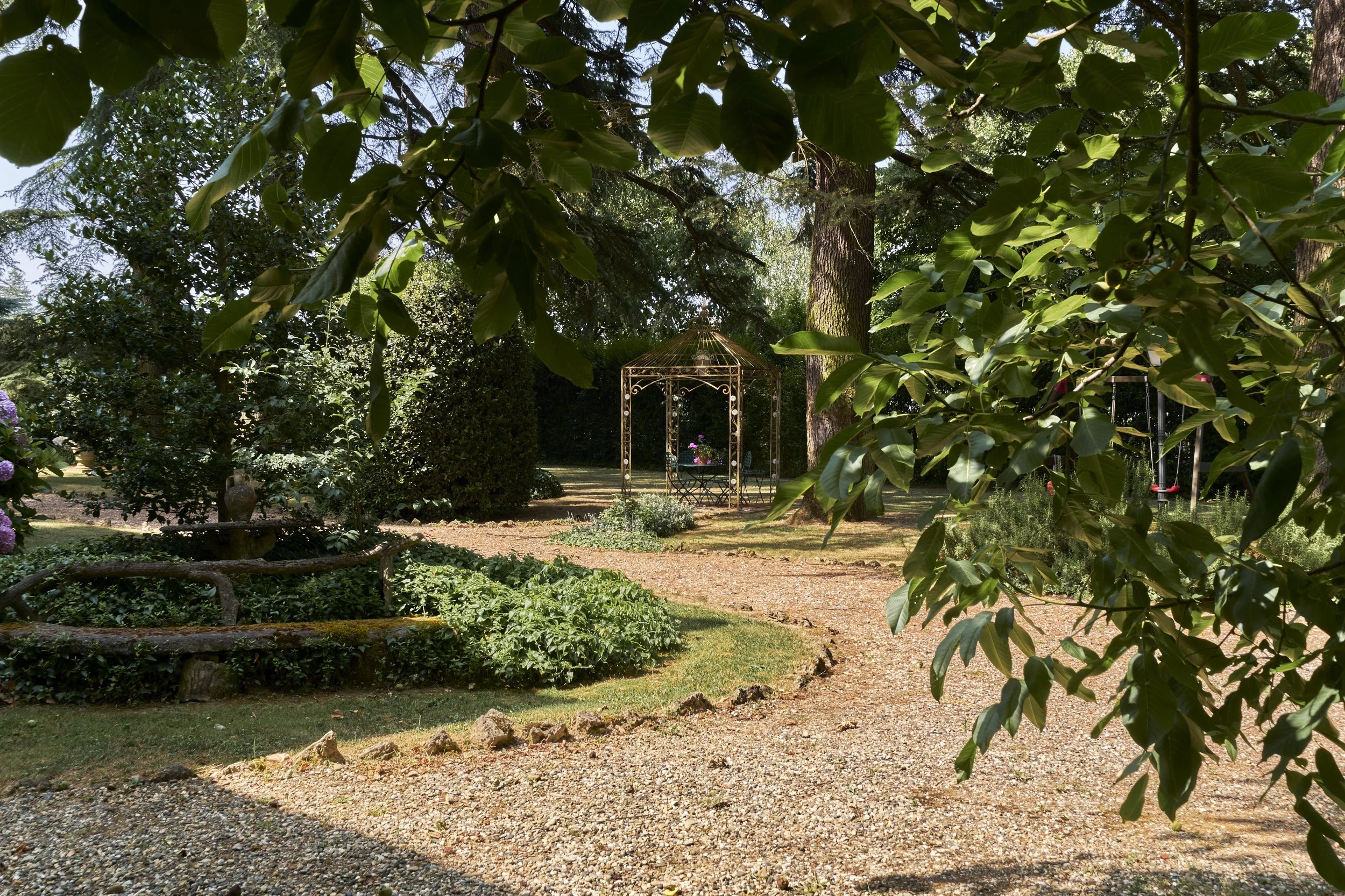 A garden with a gravel pathway, surrounded by green bushes and trees, a gazebo in the background, and a swing hanging from a tree on the right side.