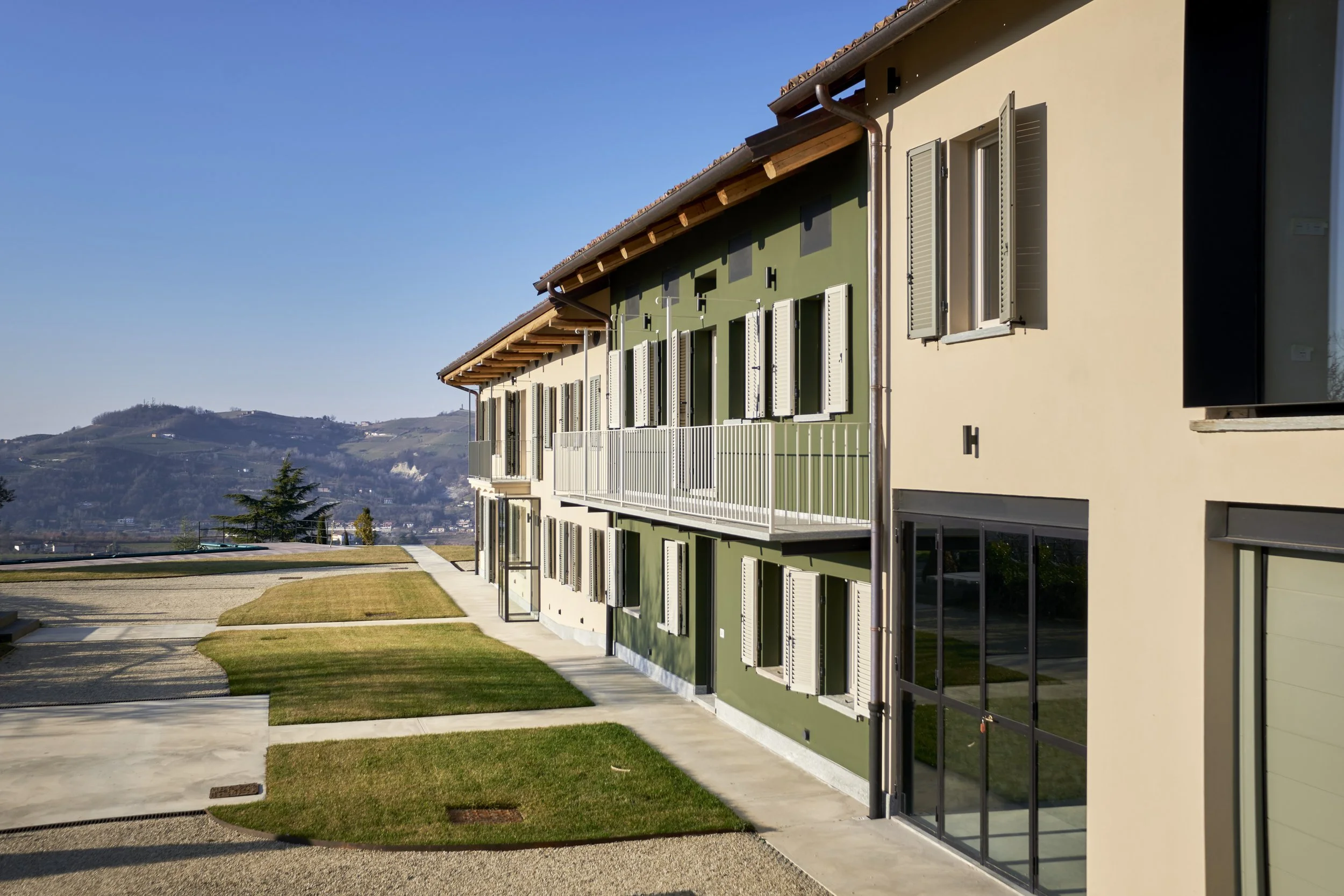 Modern residential building with green and beige exterior walls, multiple open and closed window shutters, a balcony with a white railing, and a glass door in the foreground. The building is set in a landscaped area with grass and concrete pathways,