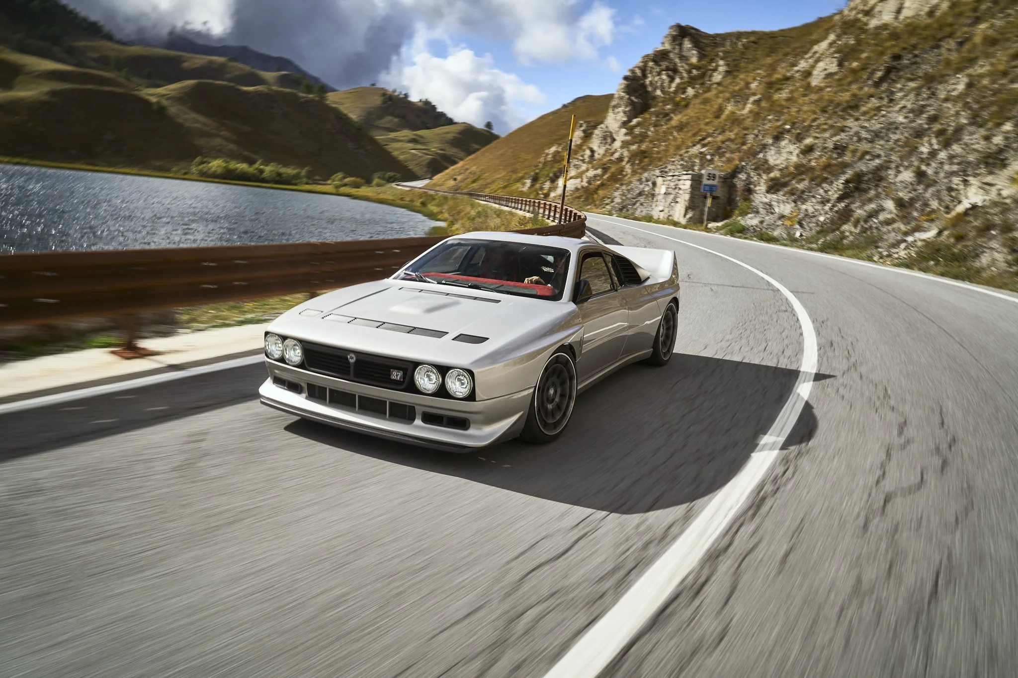 A silver vintage sports car driving on a winding mountain road alongside a body of water, with hills and rocks in the background.